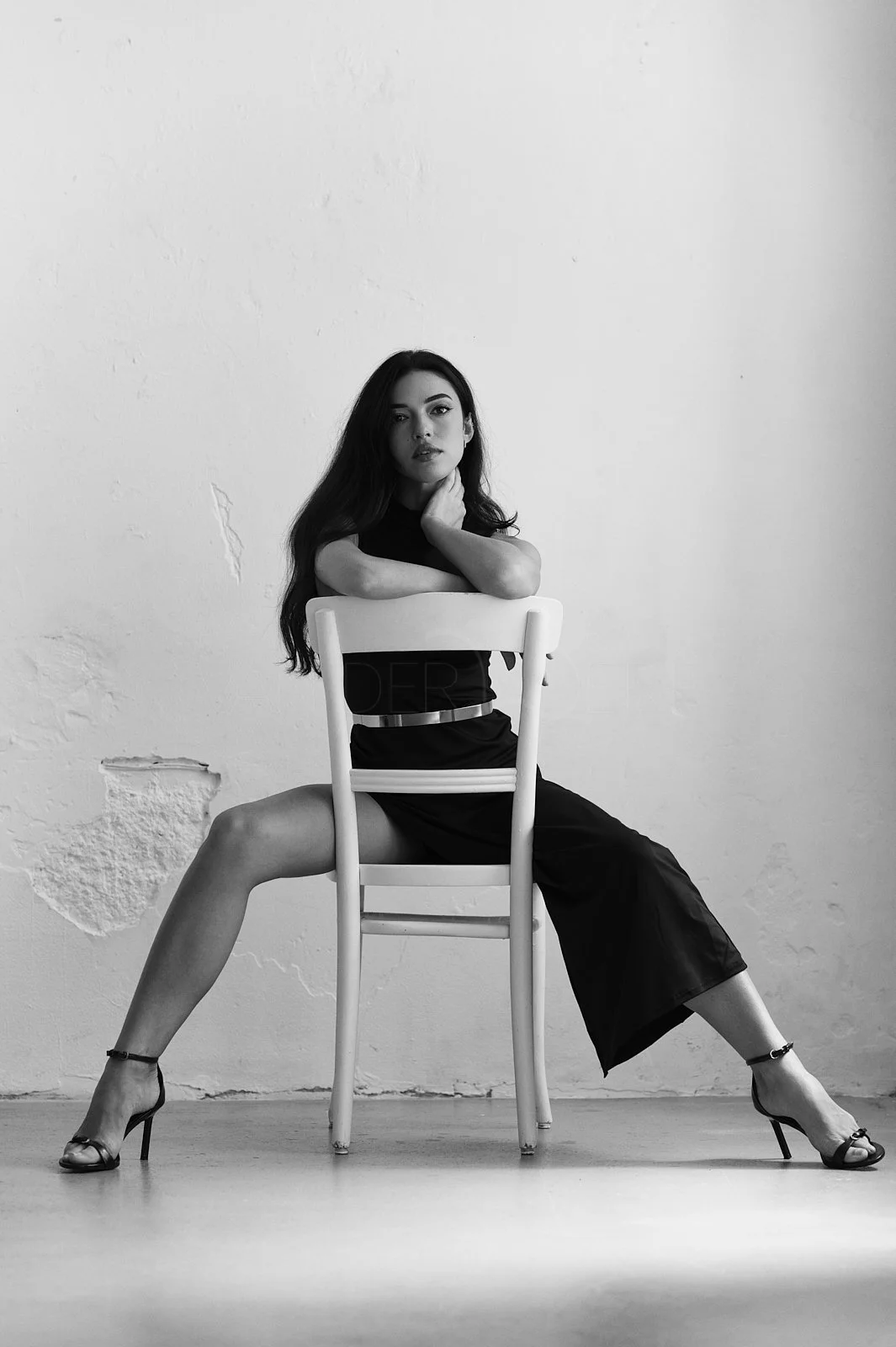 Black and white photo of a woman sitting on a chair with legs apart, one leg on the floor and the other on the seat, with her arm resting on the back of the chair and touching her face, wearing high heels and a sleeveless dress, indoors with a plain 