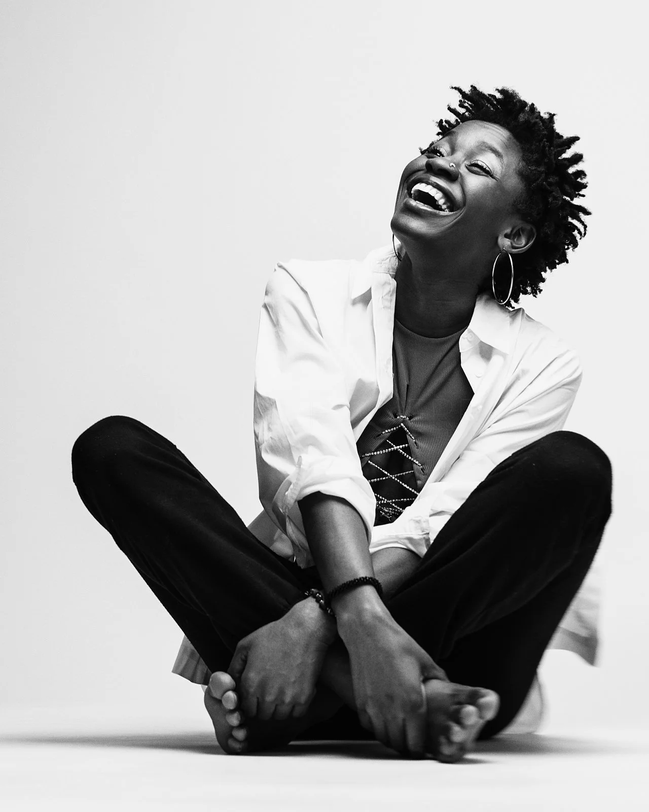 A woman with curly hair sitting cross-legged, laughing joyfully, wearing hoop earrings, a white shirt over a dark top, and black pants, in a black-and-white photograph.