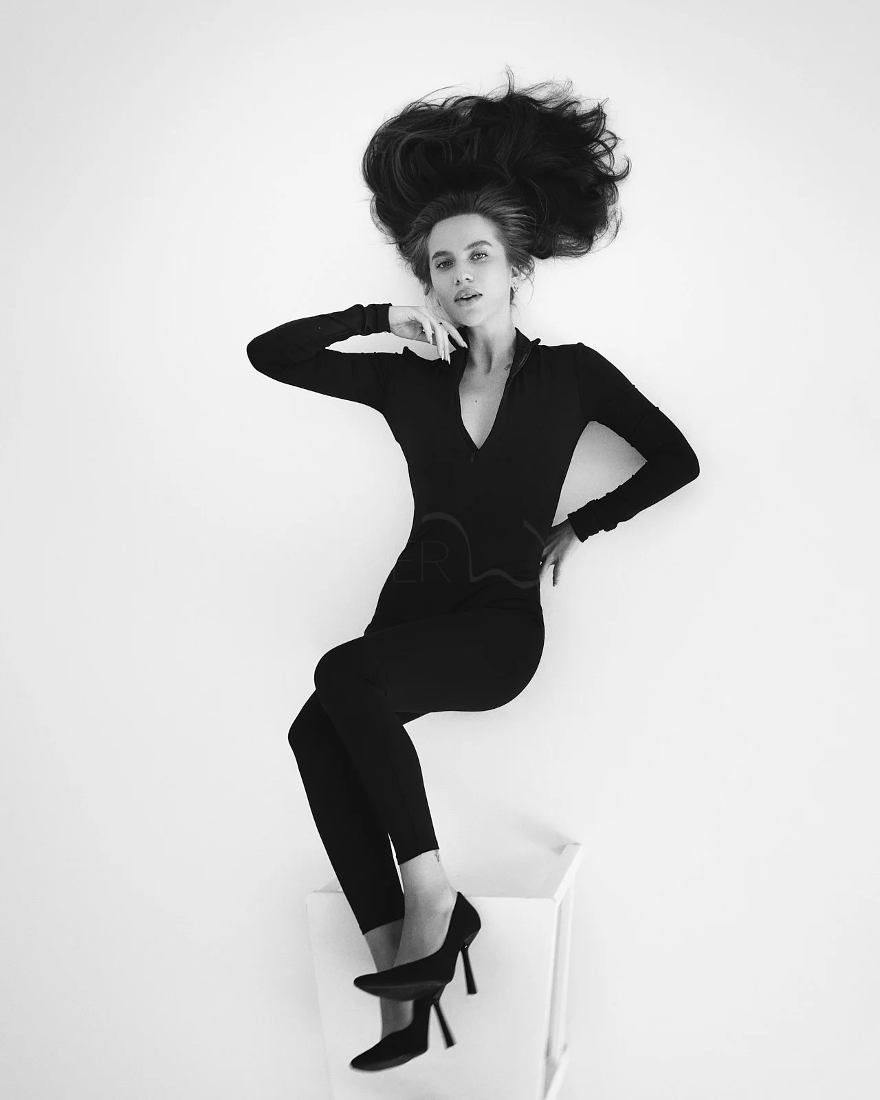 Black and white photograph of a woman with long, voluminous hair sitting on a white cube against a plain white background. She is wearing a fitted black dress and high heels, with one hand resting near her face and the other on her hip.