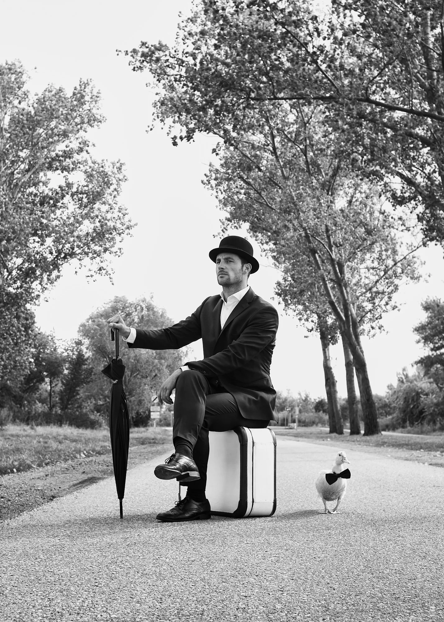 A man dressed in a suit and bowler hat sitting on a suitcase on a road, holding an umbrella, with a goose wearing a bow tie standing nearby. The scene is outdoors with trees and overcast sky in the background, in black and white.