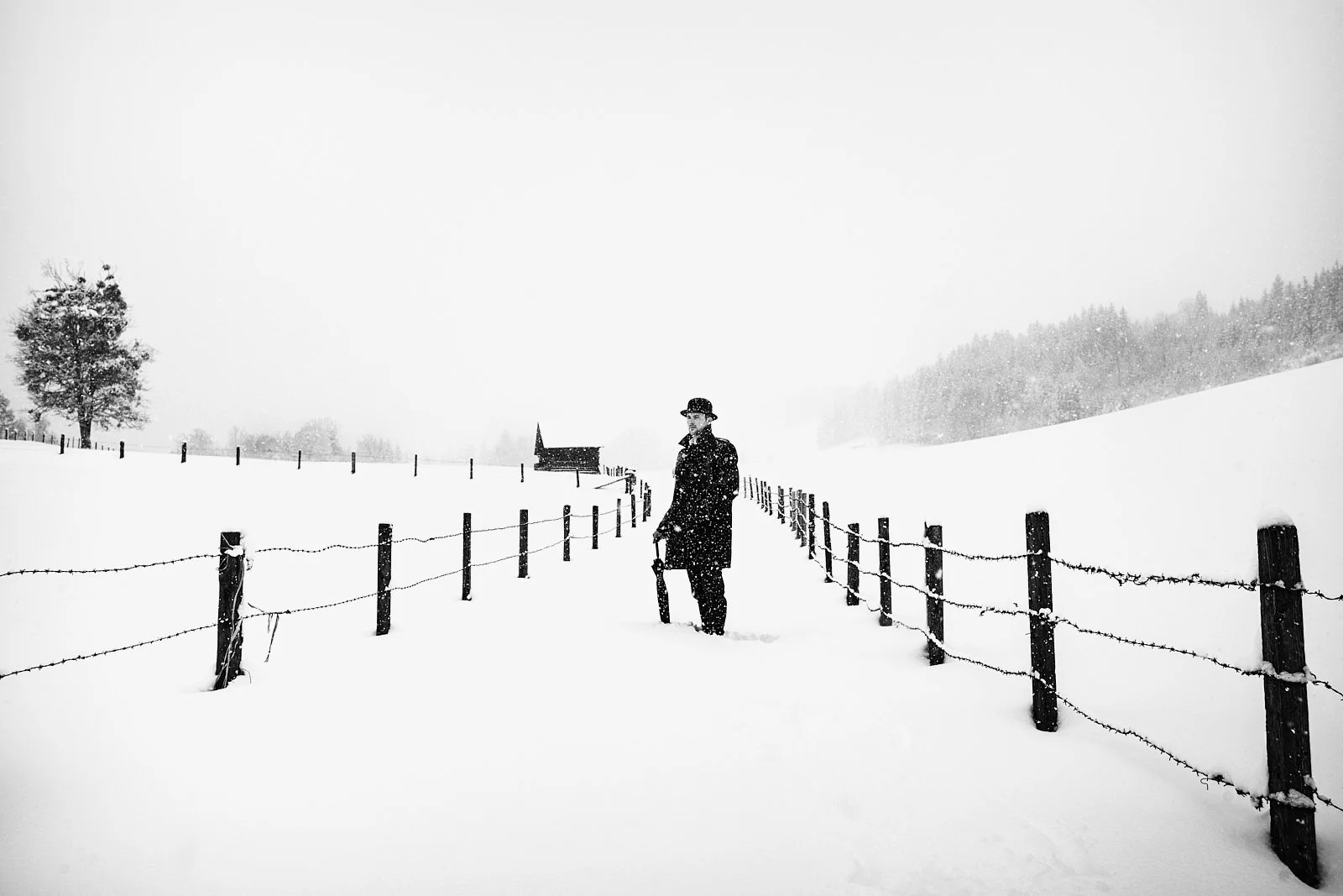 A man dressed in dark winter clothing and a hat stands in a snowy landscape, holding an umbrella, surrounded by snow-covered fences and trees, with a small building in the background.