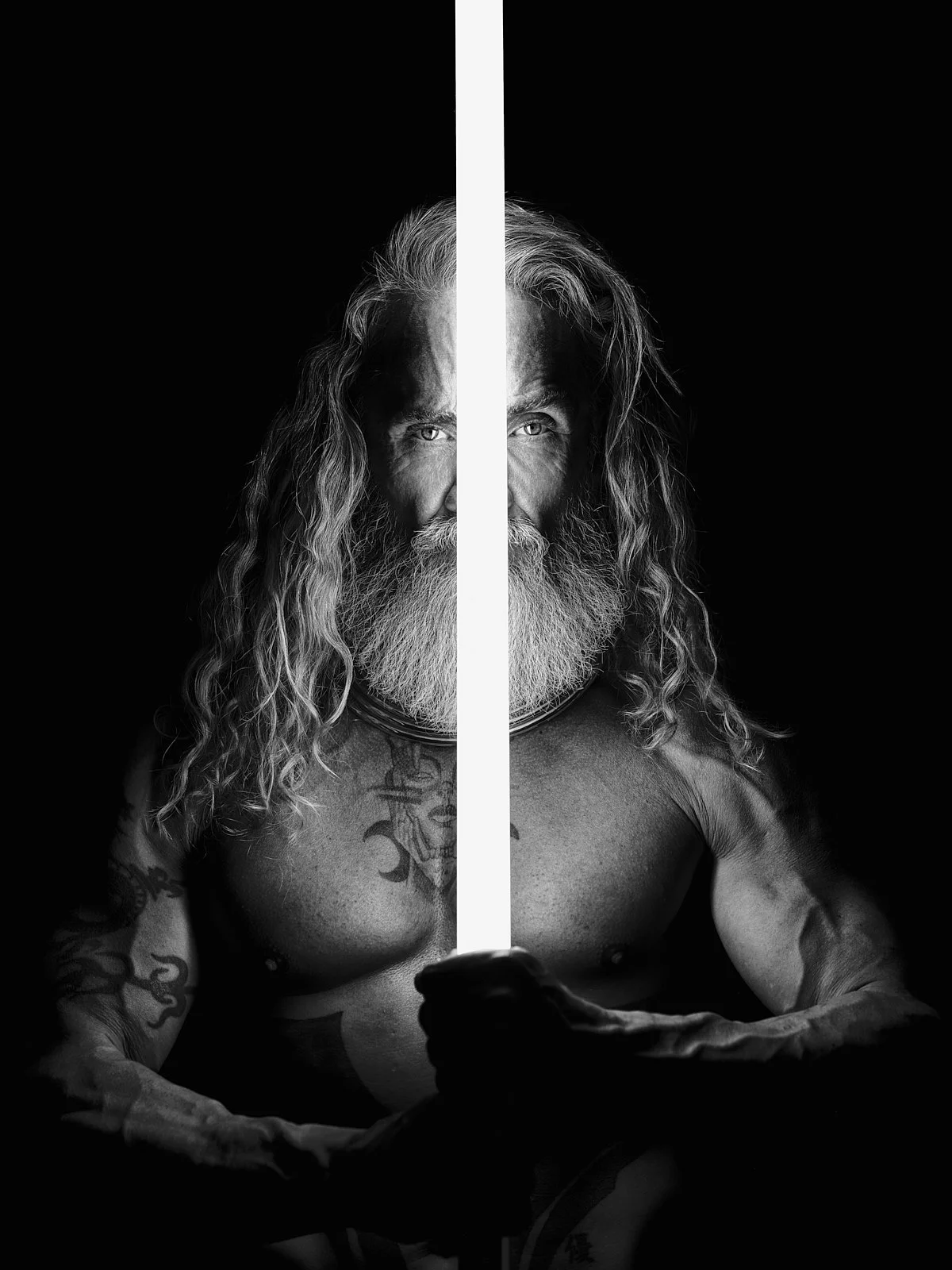 Black and white portrait of a shirtless, bearded man with long hair, holding a glowing sword with a dark background.