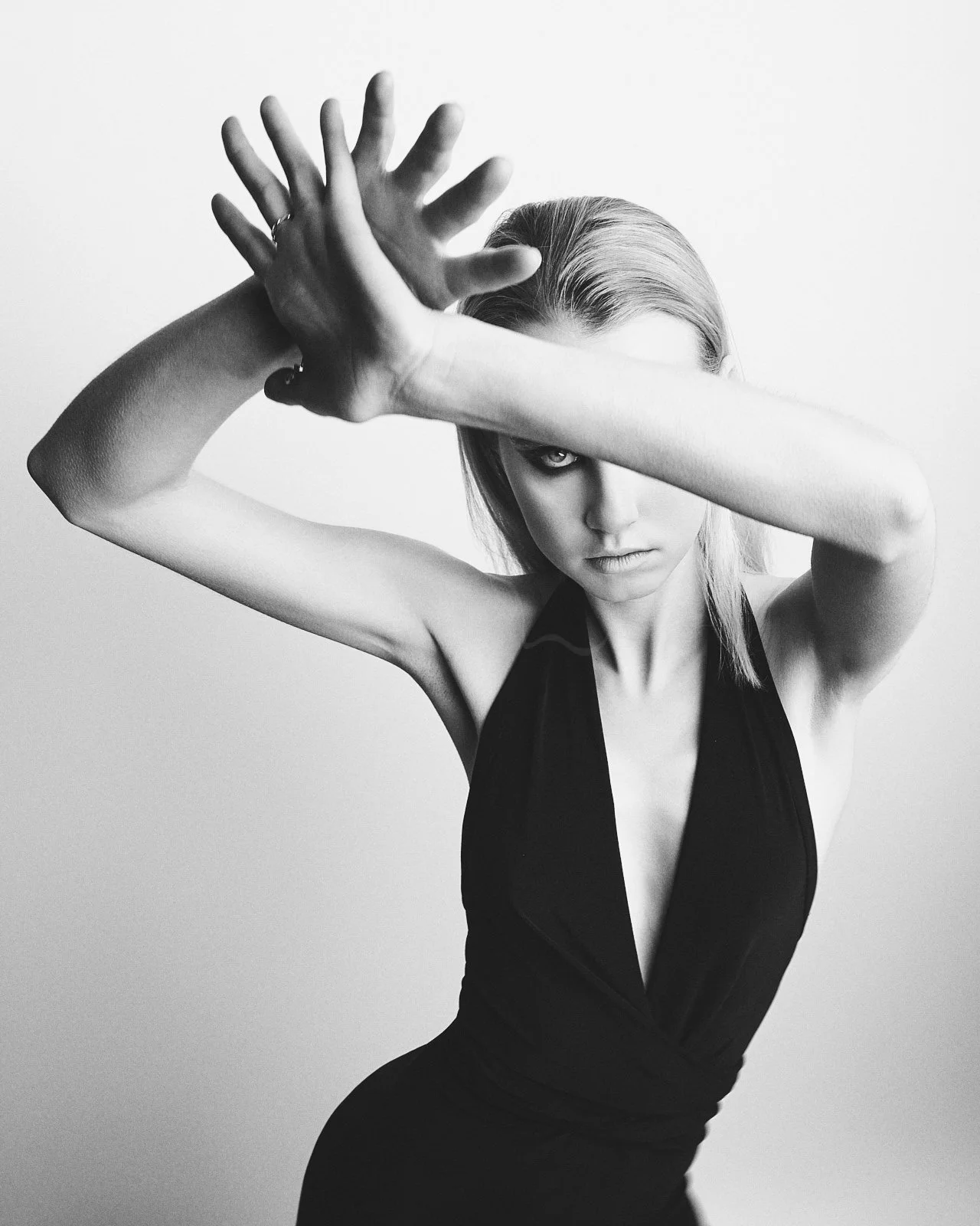A black and white photo of a woman with one arm crossed over her face, eyes intense, wearing a sleeveless dark dress, with light-colored hair.