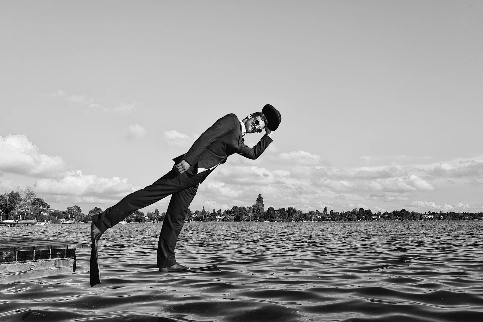 Black and white photo of a man in a suit, sunglasses, and a hat balancing on a surfboard in water, holding his hat