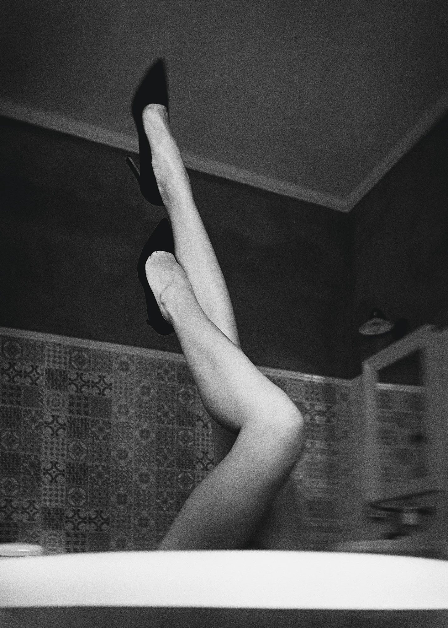 Black and white photo of two legs extended upward, wearing high-heeled shoes, with a tiled wall and ceiling in the background.