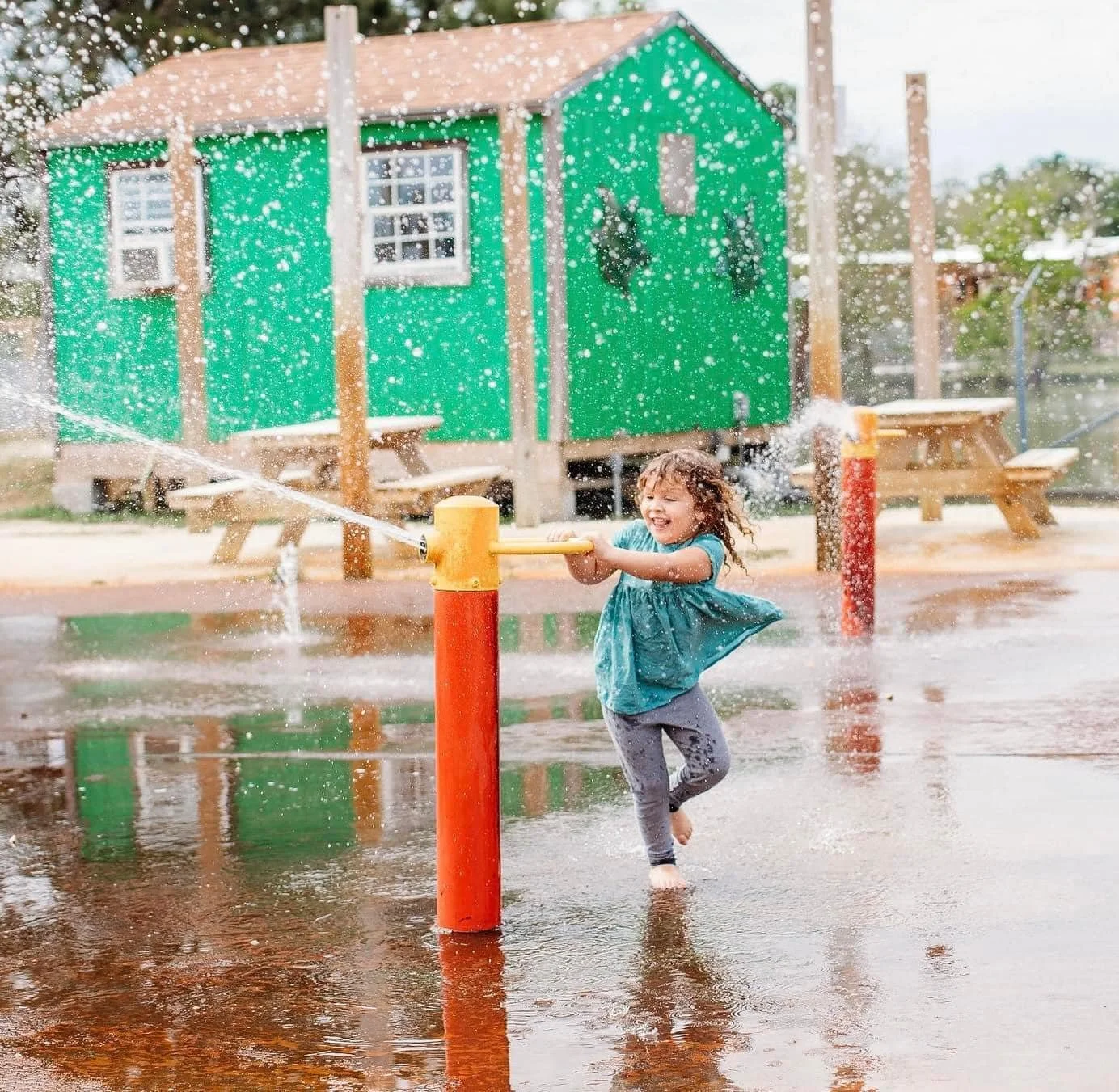 A young girl in a blue shirt and gray leggings playing with a water hose at a spray park, splashing and smiling with her arms around the hose, with a green building and picnic tables in the background.