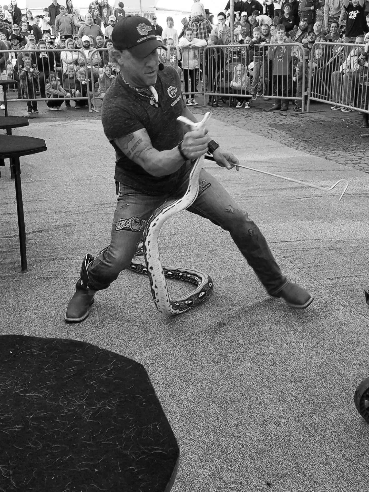 A man performing a snake charming act by holding a snake with one hand and a hook with the other in front of a crowd at an outdoor event.