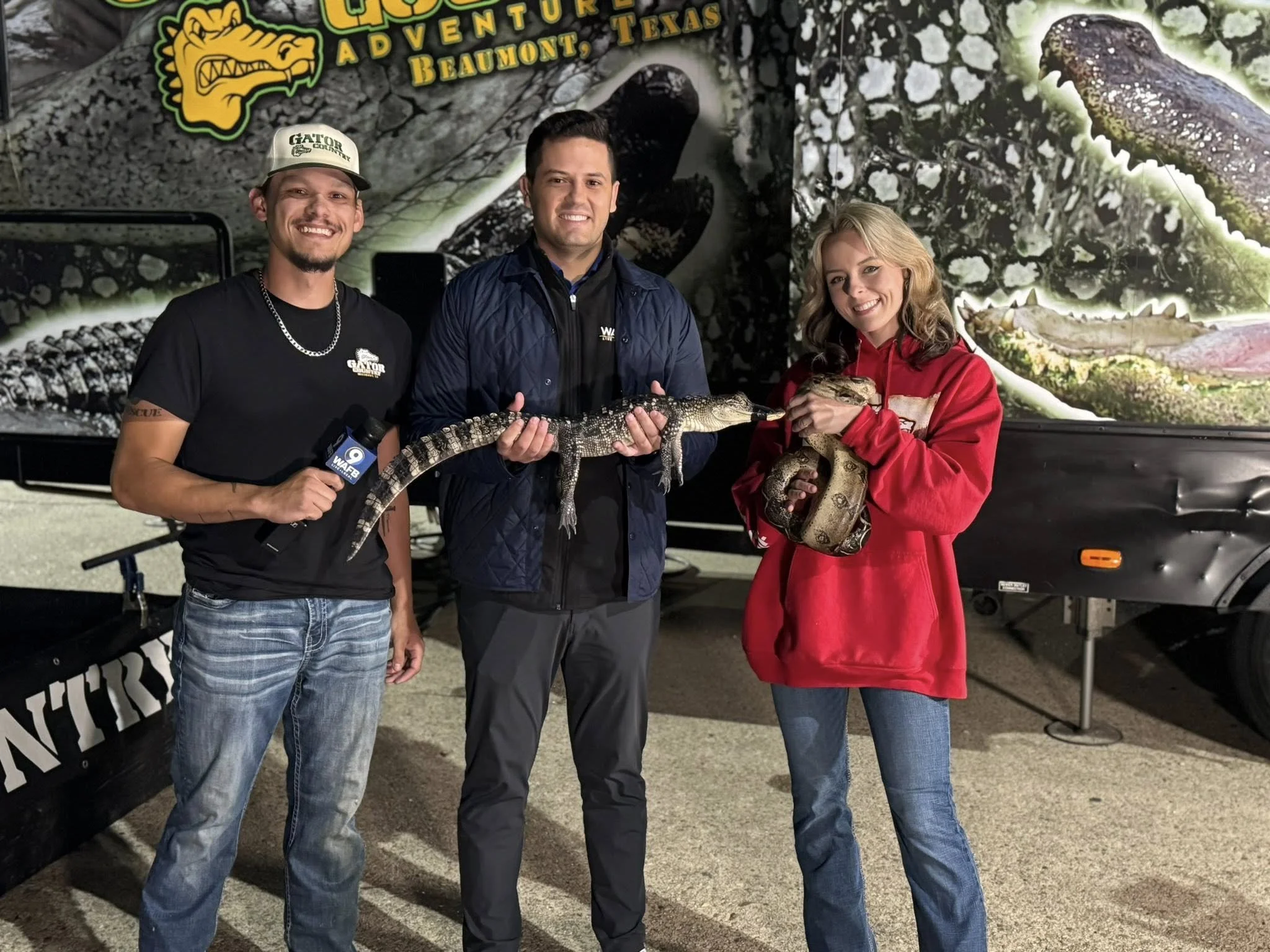 Three people are standing in front of a promotional backdrop for Gator Country Adventure in Beaumont, Texas. The person on the left is holding a microphone and smiling, wearing a black t-shirt, a cap, and jeans. The person in the middle is holding a small alligator and smiling, wearing a dark jacket and pants. The person on the right is holding a larger alligator and smiling, wearing a red hoodie and jeans.