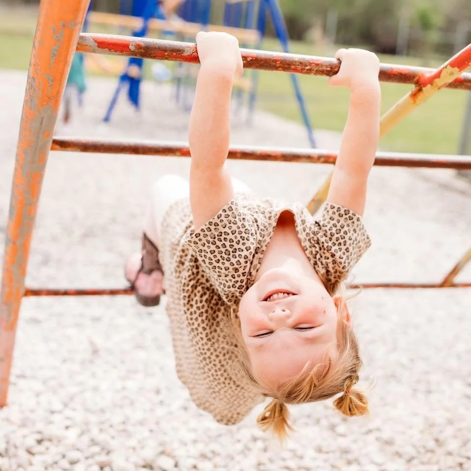 A young girl hanging upside down on a playground jungle gym, smiling with her eyes closed, wearing a leopard print shirt and pink pants.