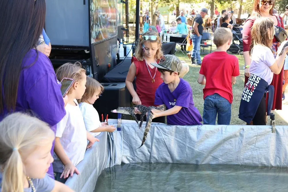 Children and adults observing a small crocodile or alligator held by a boy in a purple shirt and camouflage hat at an outdoor event.