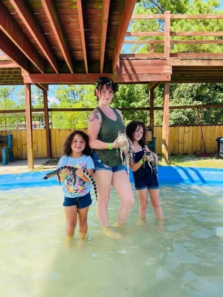 Three women stand in a shallow pool holding small crocodiles or alligators. One young girl on the left holds a young crocodile, while the woman in the middle holds a larger crocodile. The third woman on the right is holding a smaller crocodile. The background shows a backyard with a wooden deck and trees.