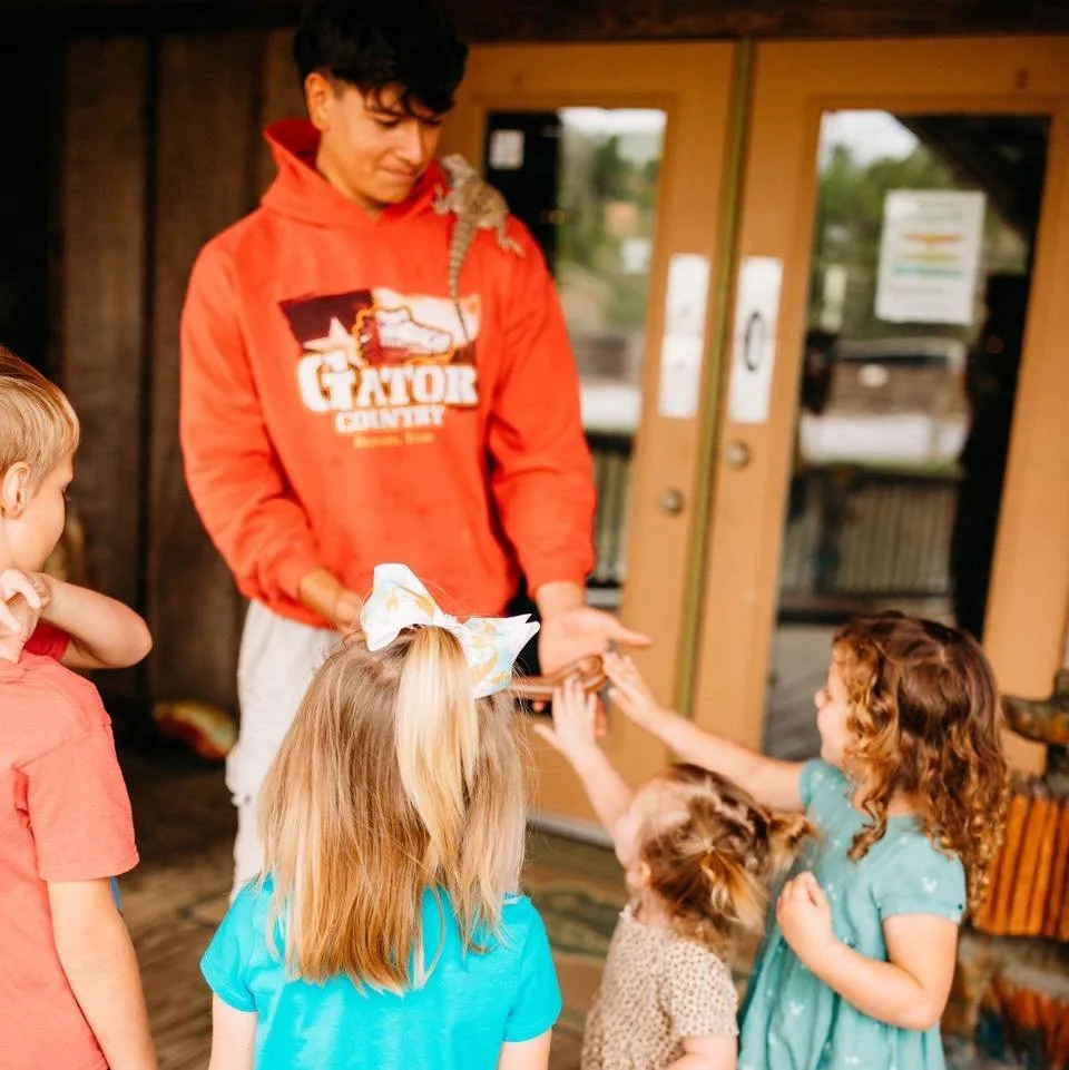 A young man wearing a red hoodie with a lizard on his shoulder and a group of children inside a wooden building, with some children reaching out and touching his hand.