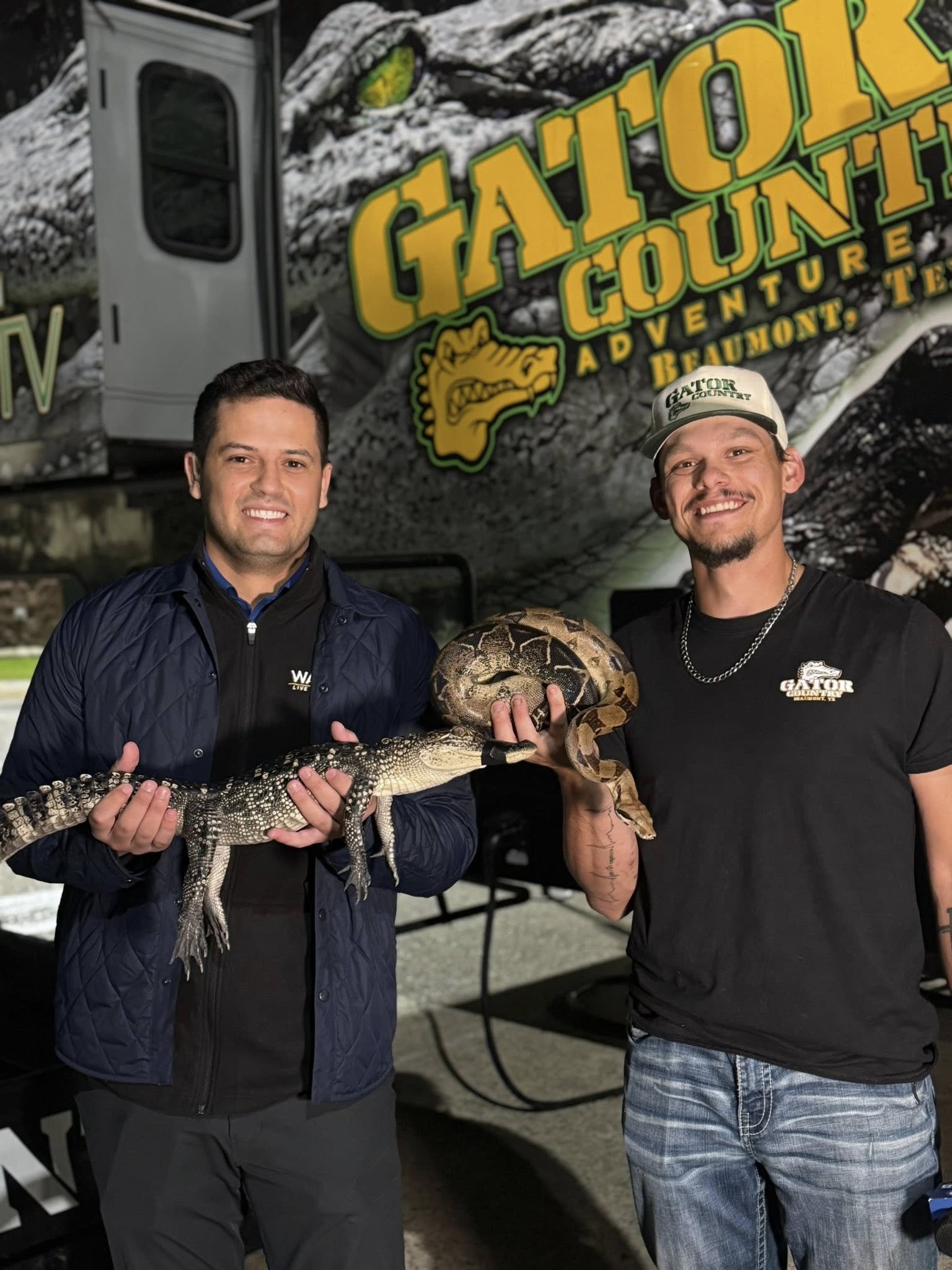 Two men standing outside in front of a large Gator County Adventure Tour sign, holding a small alligator and a snake.