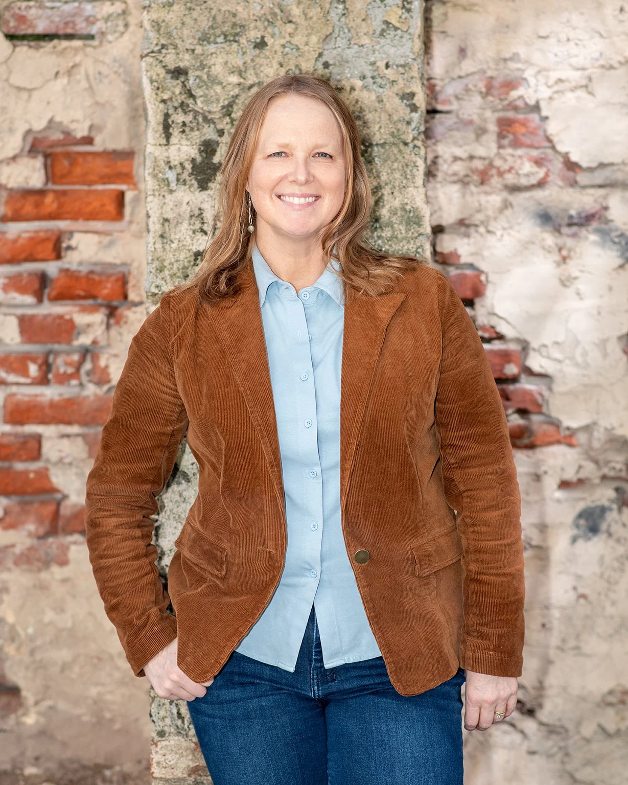 A woman standing outdoors in front of old brick and stone wall, smiling, wearing a brown blazer, light blue shirt, and blue jeans.