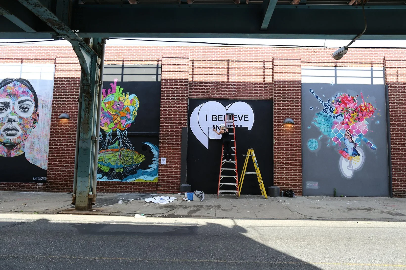 A person on a ladder painting a mural with a large heart that has the words "I BELIEVE IN YOU" inside on a brick wall. There are three other colorful murals on the wall, including a woman's face, an abstract fish scene, and a bird in flowers.