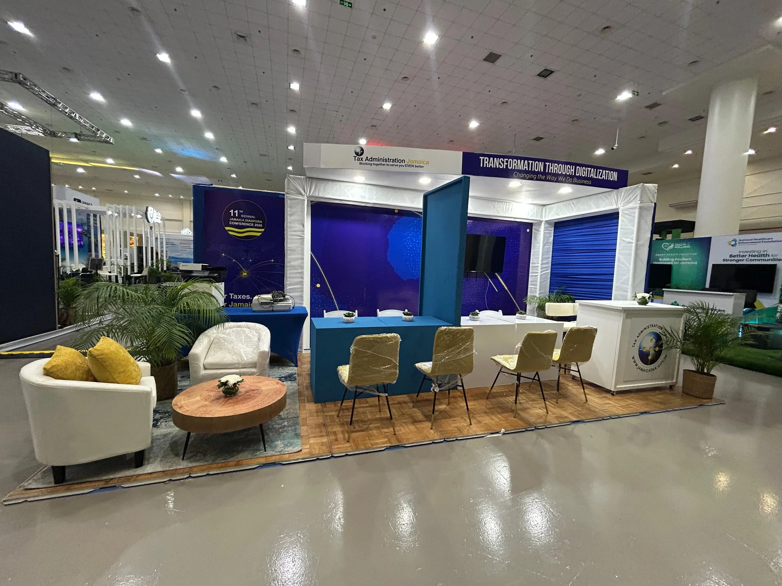 An indoor exhibition booth for the Jamaica Tax Administration with a large sign reading "Transformation Through Digitalization" and seating arrangements, plants, and displays related to Jamaica tax services.