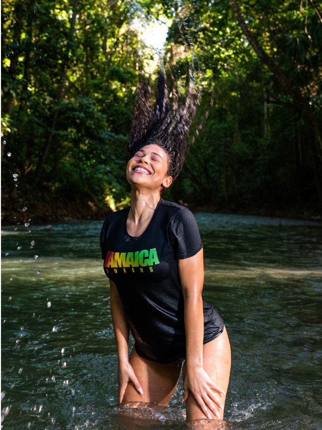 Woman standing in a river, hair flipped back, smiling with eyes closed, wearing a black T-shirt with 'Jamaica Queens' in colorful letters, surrounded by greenery.
