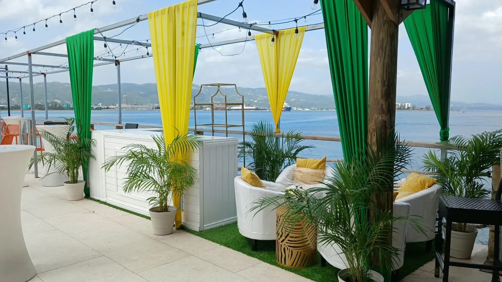 Outdoor waterfront lounge area with white furniture, yellow and green curtains, potted plants, and string lights, overlooking a body of water with hills in the background. Designed by MCM Collective Jamaica.