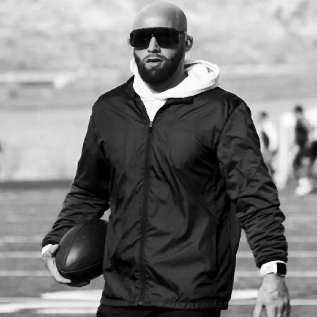 A man holding a football on a sports field, wearing black sunglasses, a black jacket, and a white hoodie underneath.