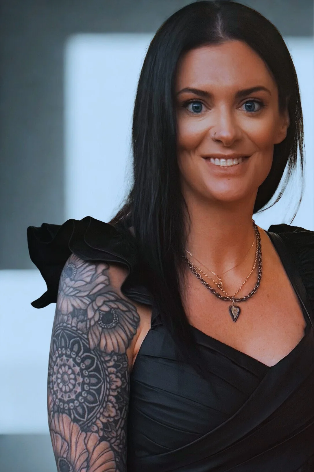 Laura Beth Burkhalter or LB Burkhalter with long black hair, blue eyes, and a tattoo on her arm, wearing a black dress and layered necklaces, smiling at the camera about women's sober living in Colorado.