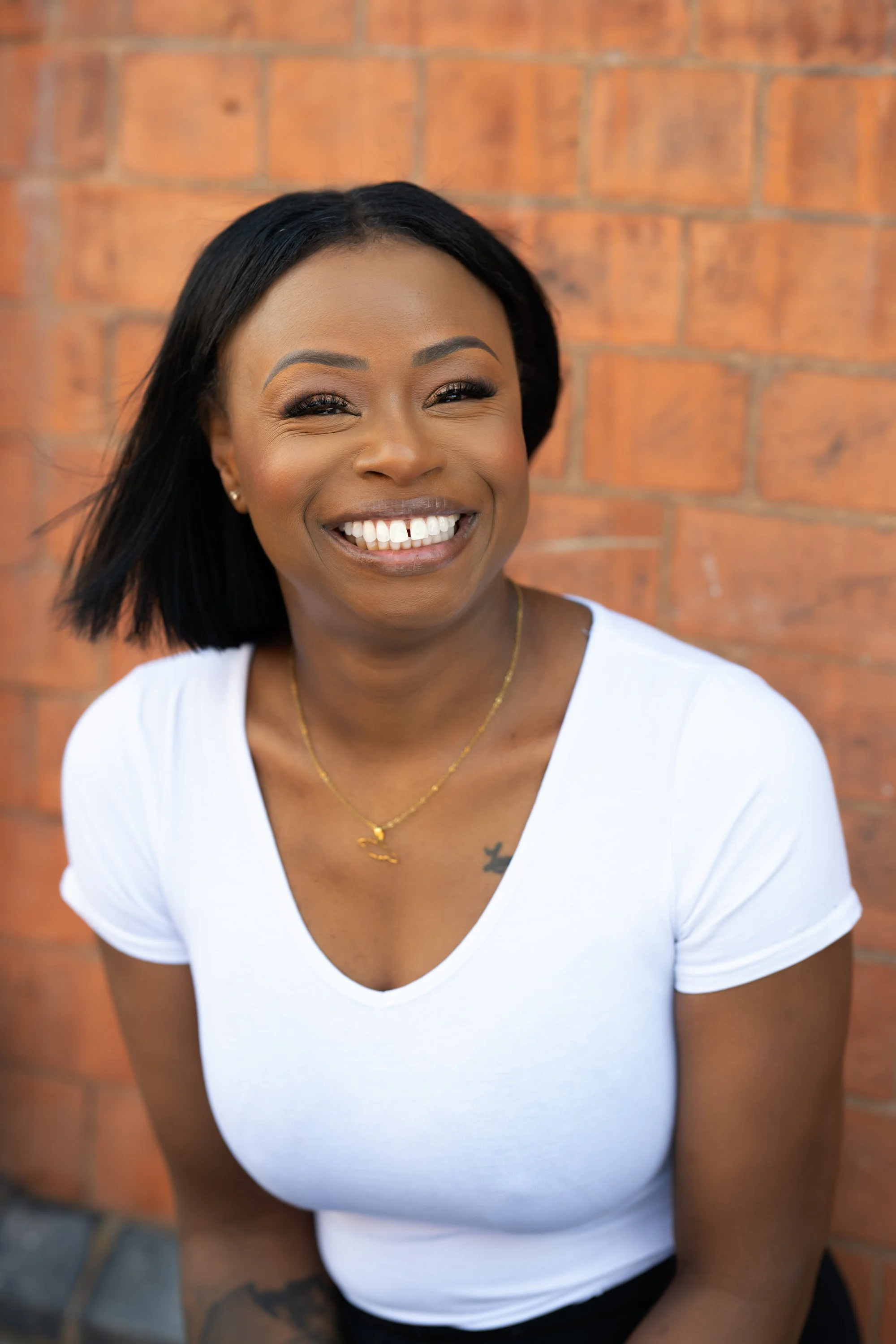 A smiling Black woman wearing a white V neck top and a gold necklace with the outline of Jamaica as a pendant is standing in front of a brick wall.