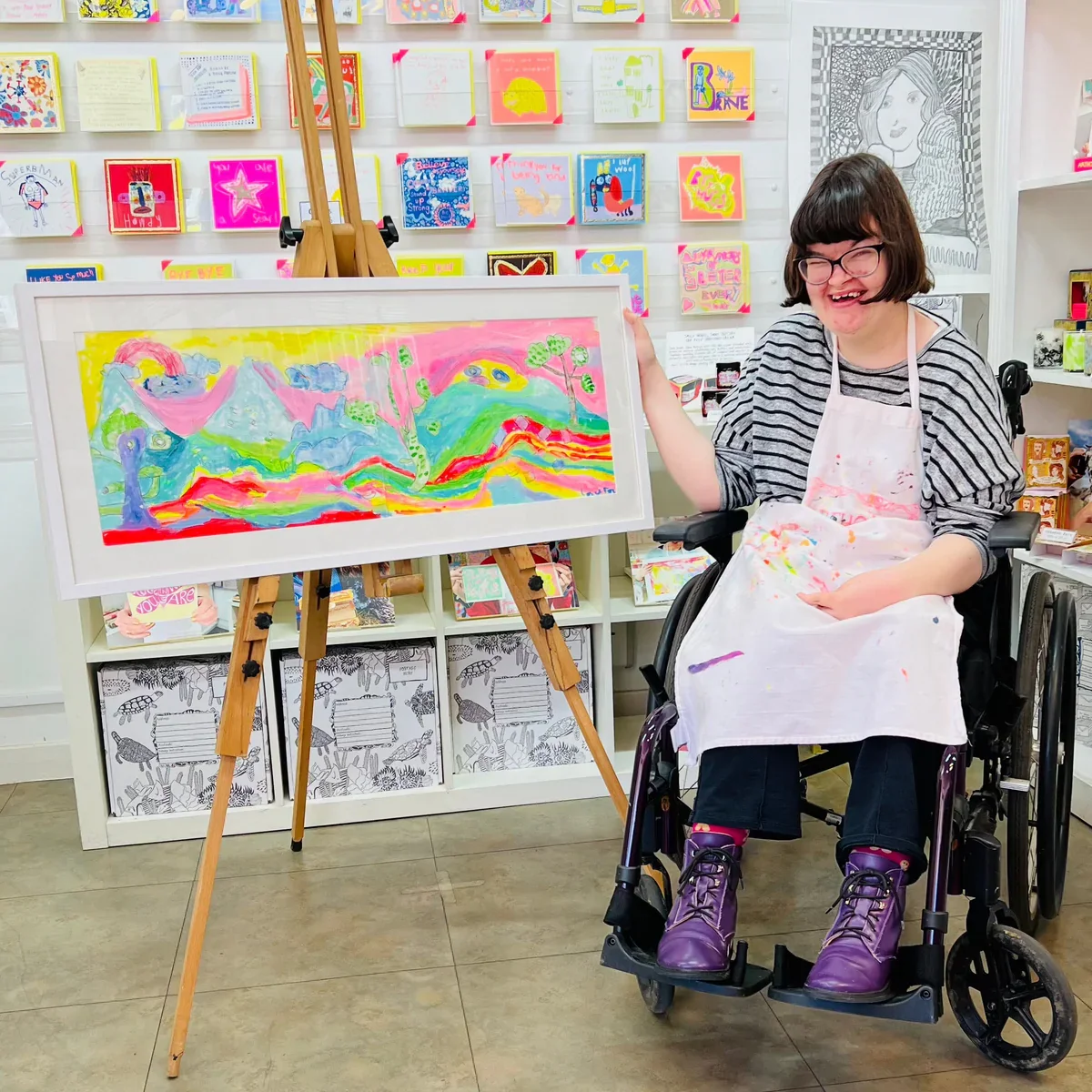 Young woman in a wheelchair wearing a striped top, purple boots, and a paint-splattered apron, smiling next to her colorful landscape painting on an easel, in an art gallery with decorated walls.