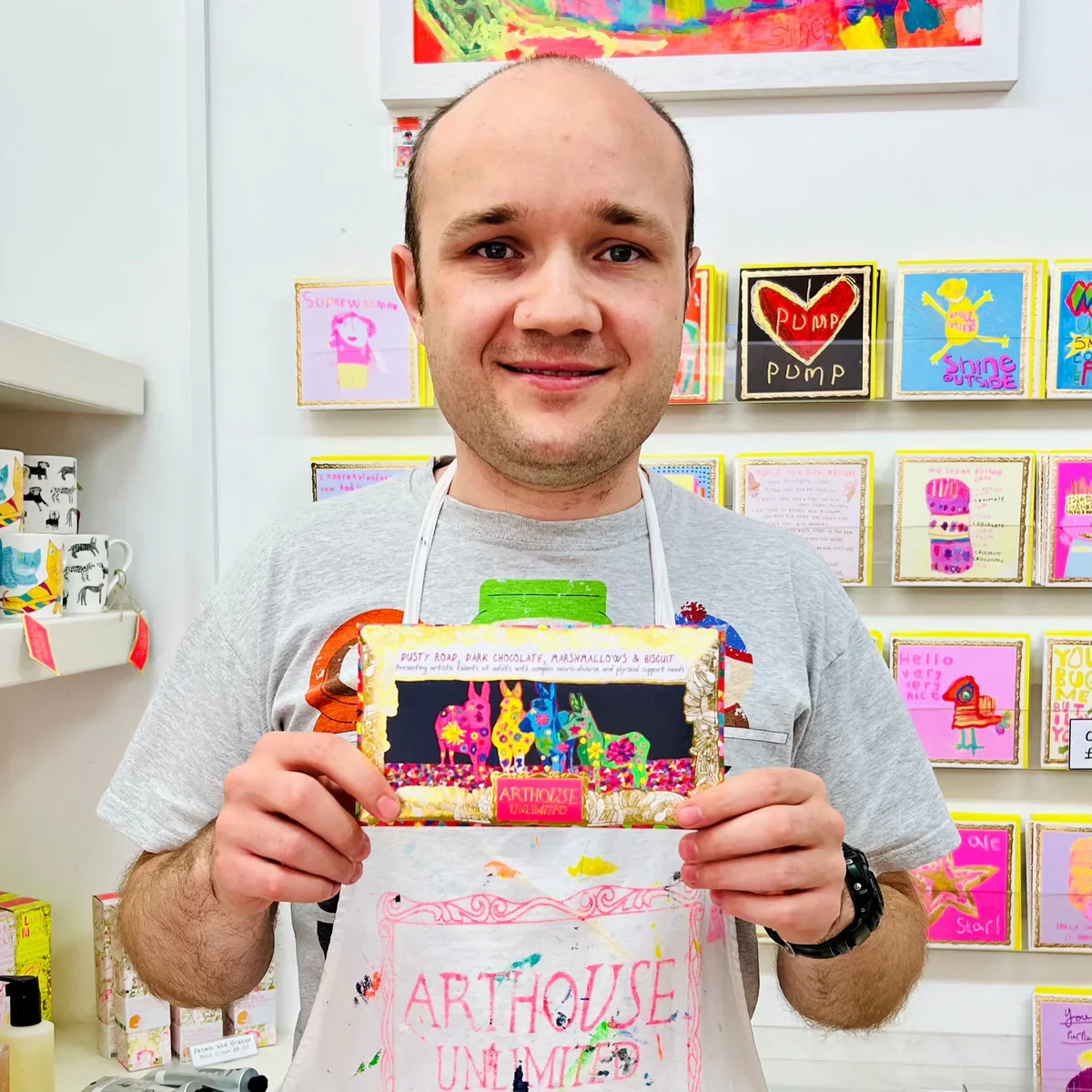 A man with a bald head and smiling face holding a colorful artistic card at an art store. The card has bright, multicolored illustrations of animals and is labeled 'ARTHOUSE UNLIMITED.' The man is wearing a casual gray t-shirt and a white apron with 