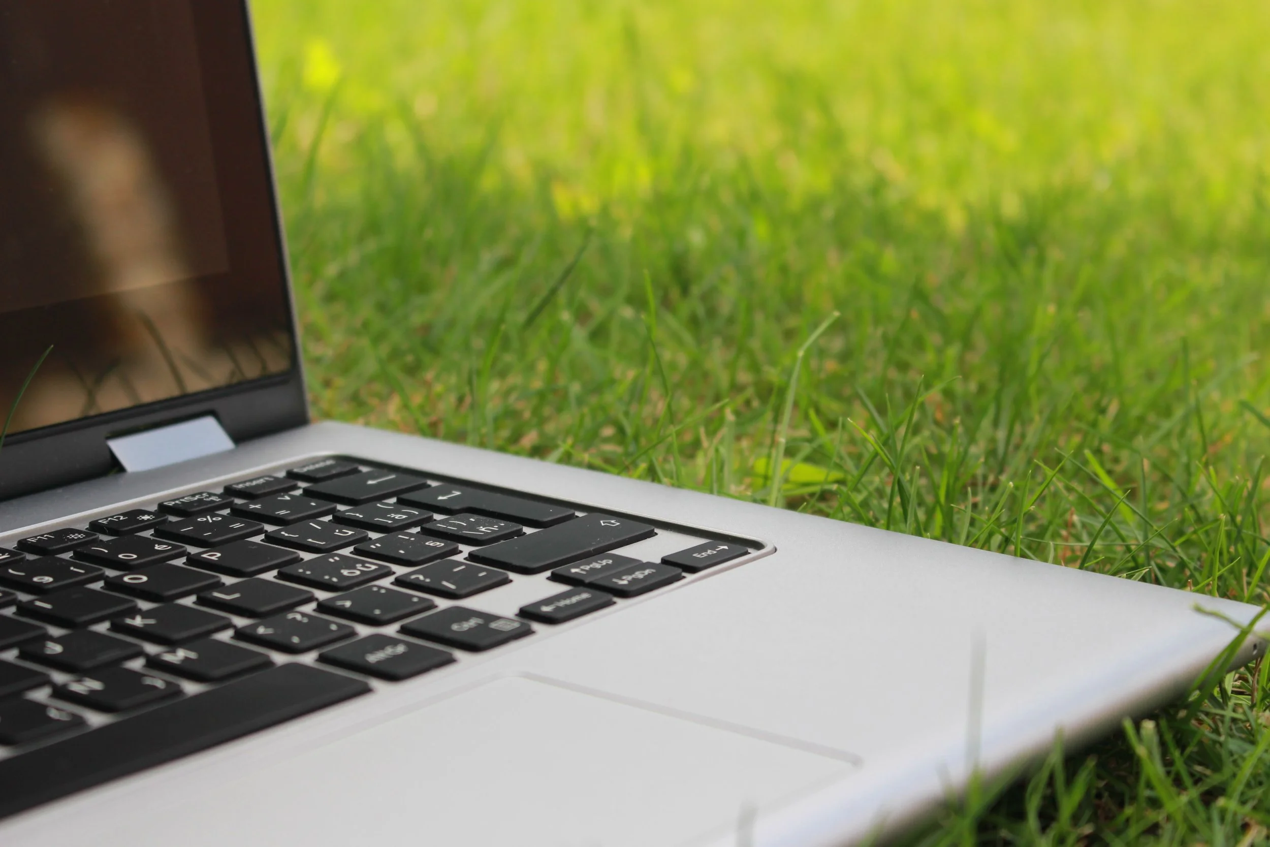 Close-up of a laptop keyboard on grass with a blurred outdoor background.