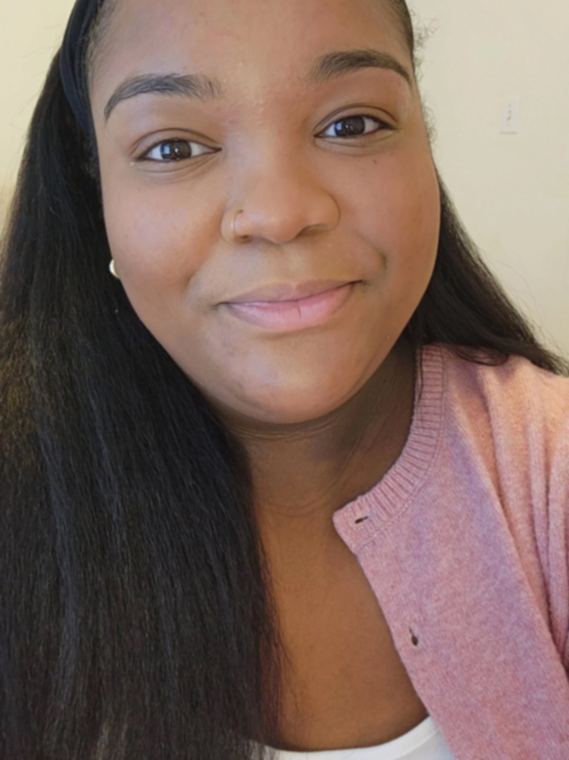 A young woman with medium brown skin, long black hair, and brown eyes smiling at the camera, wearing a pink cardigan over a white top, and a black headband.