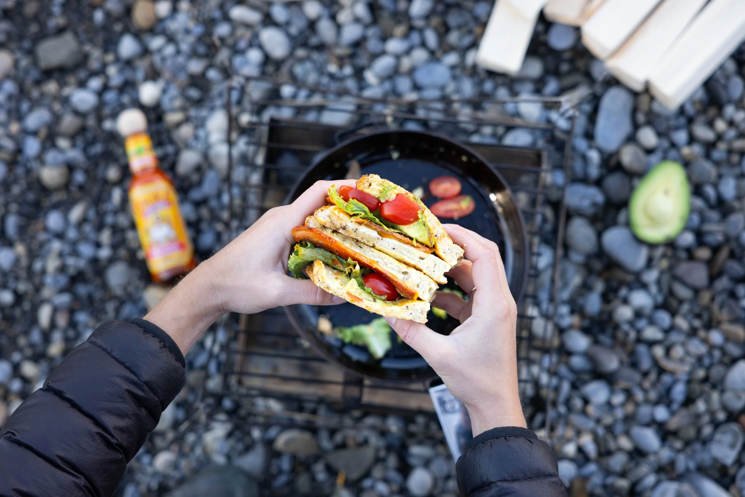 hands holding breakfast sandwich with bacon, tomato, egg, and avocado