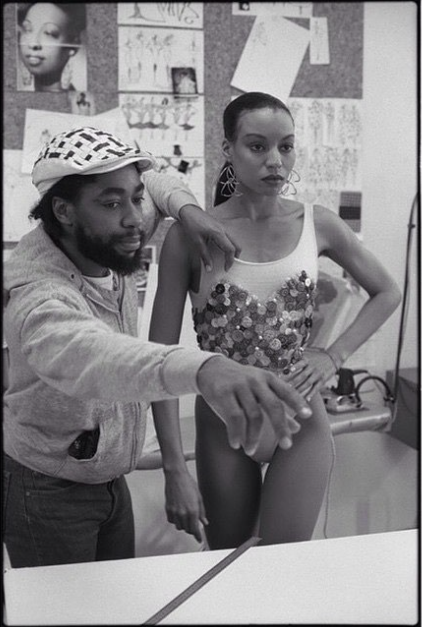 A man and a woman are posing in front of a bulletin board with various posters and images. The man is wearing a patterned cap and hoodie, and the woman is wearing large earrings and a sequined top. The man is pointing towards the woman, and they are 