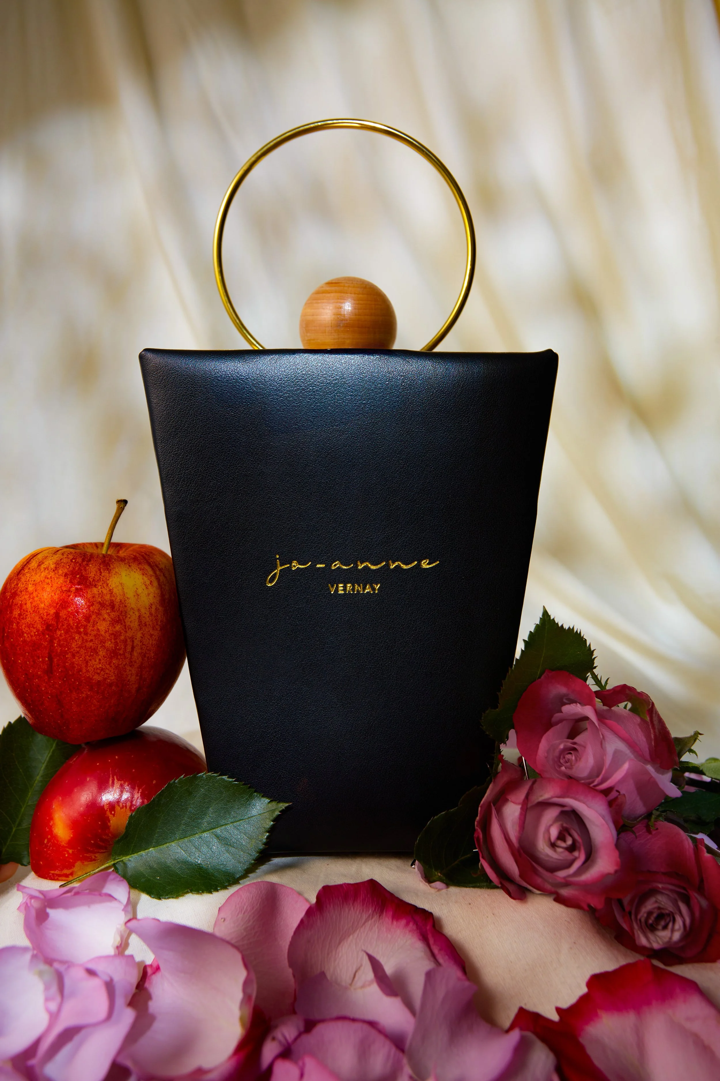 A black book with gold lettering that reads "joanne VERNY" is surrounded by pink roses with green leaves, red apples, a wooden ball, a gold ring, and a beige fabric background.