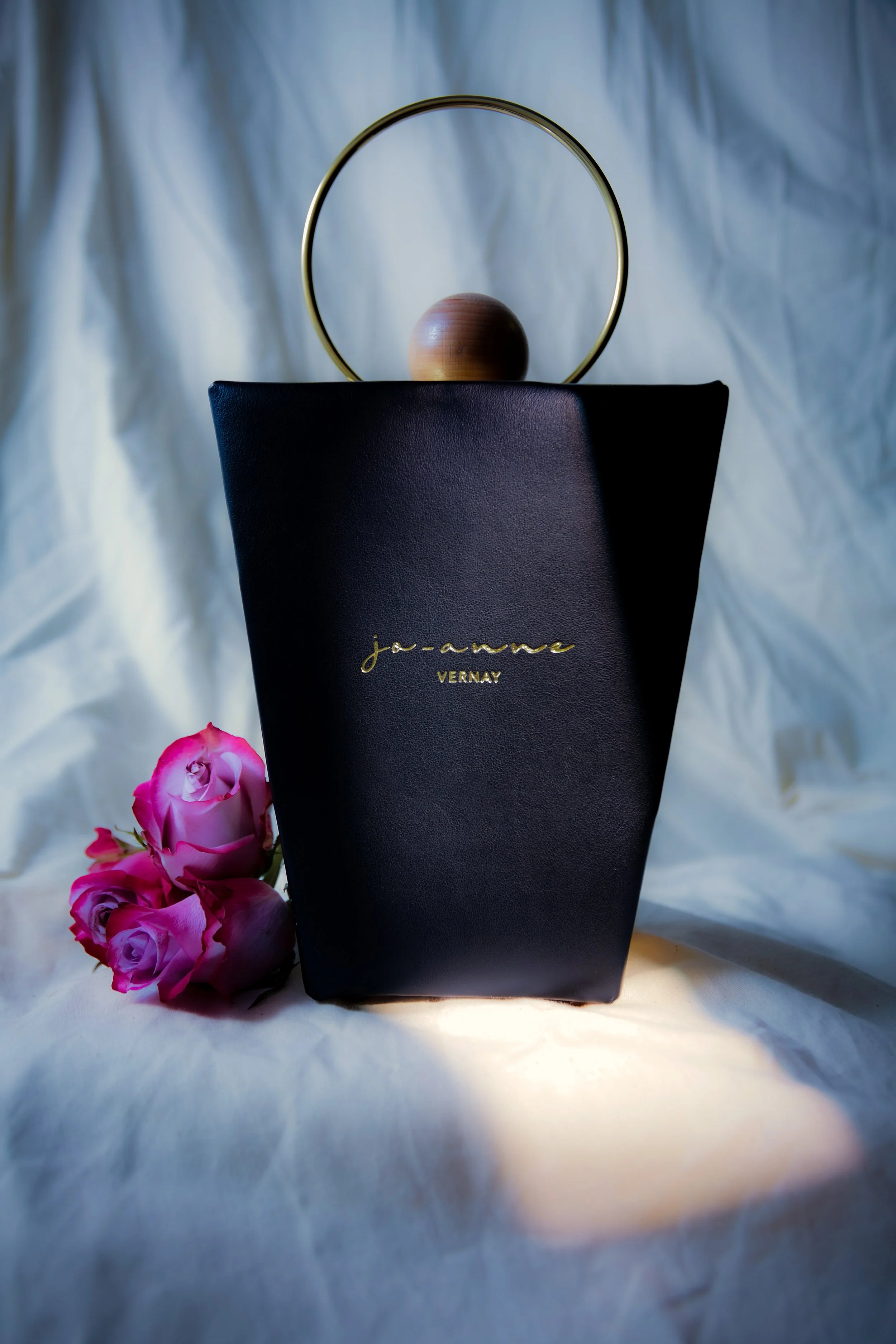 A black designer handbag with gold lettering placed on a white fabric background, with pink roses in the foreground and a gold circular decorative piece behind it.