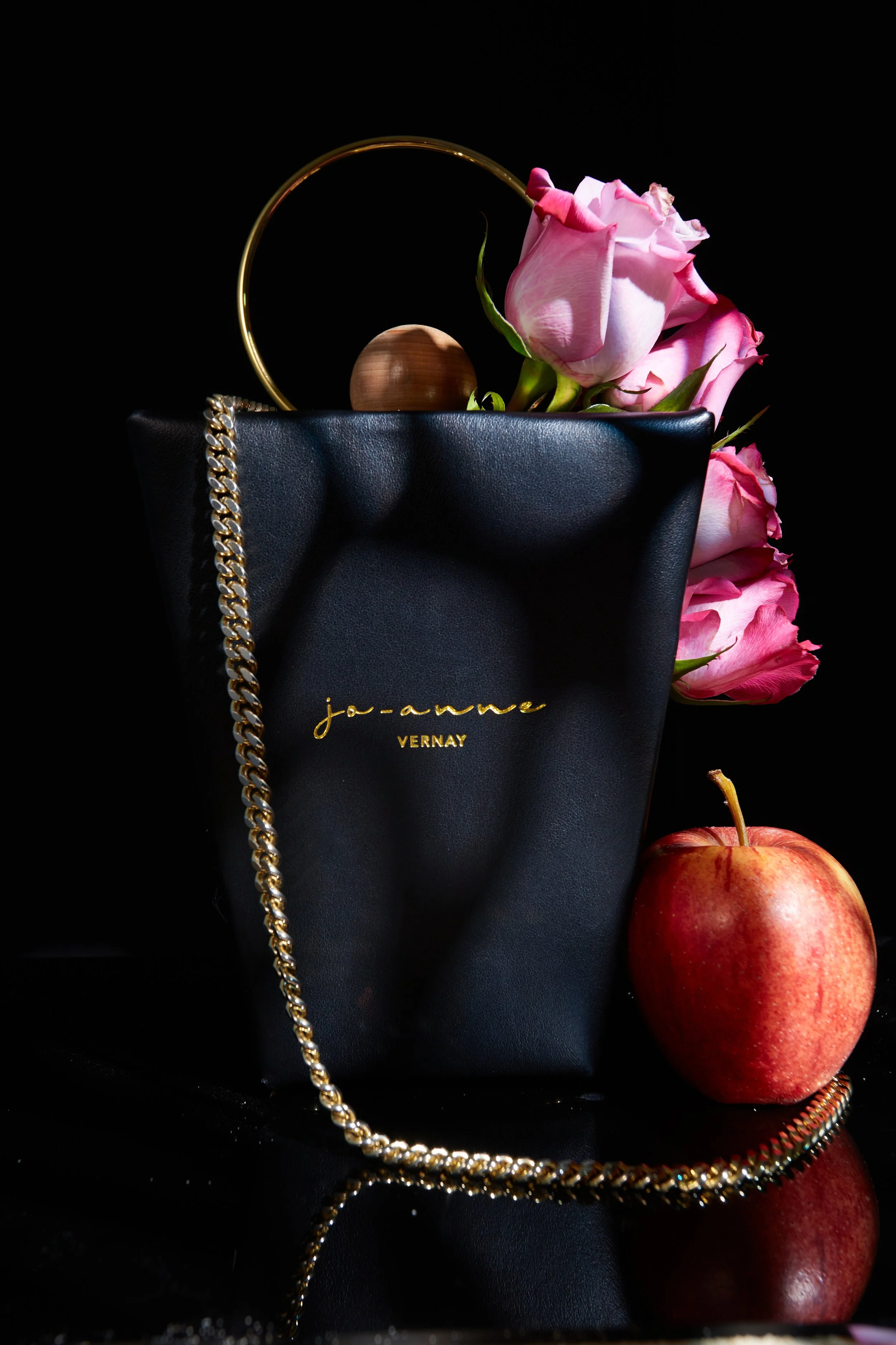 A black handbag with gold lettering and chain strap, holding pink roses, set against a dark background with an apple nearby.