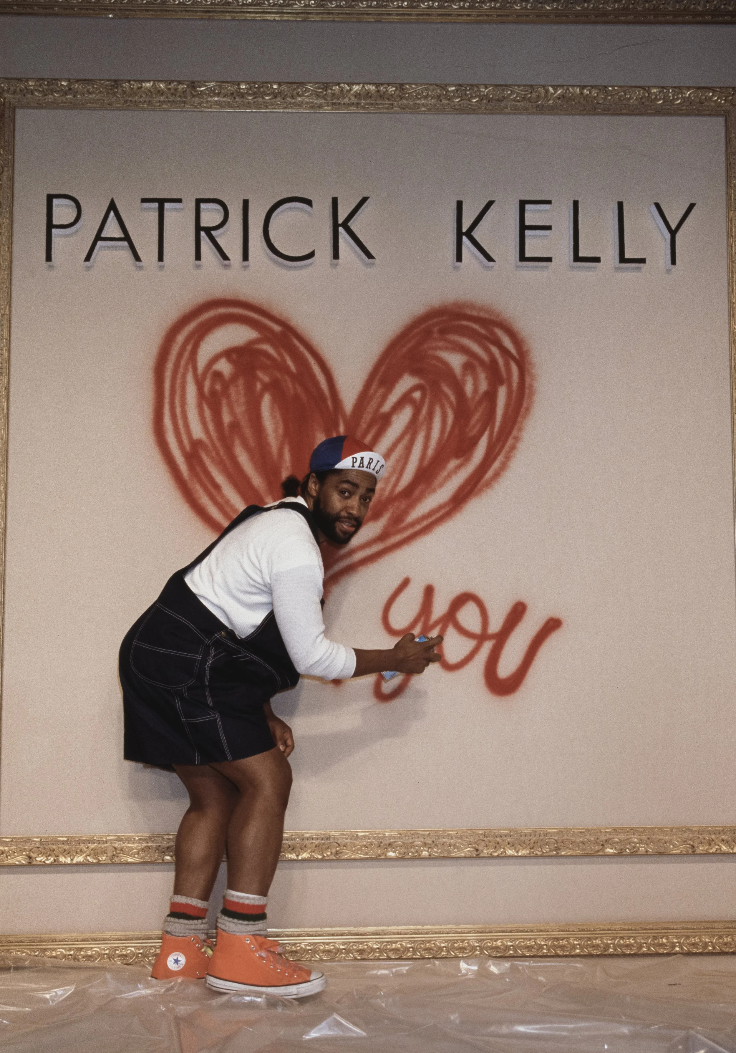 A person wearing orange oversized Converse sneakers, striped socks, and a white shirt with black shorts is standing in front of a wall sign. The sign displays the name 'Patrick Kelly' and features a large red heart drawing with the graffiti word 'You