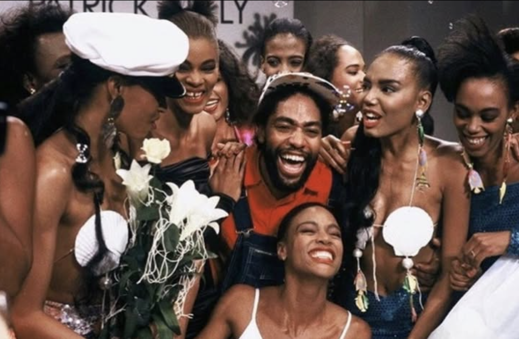 A group of diverse women and one man smiling and celebrating, dressed in colorful and festive clothing with floral accessories, in a joyful moment.