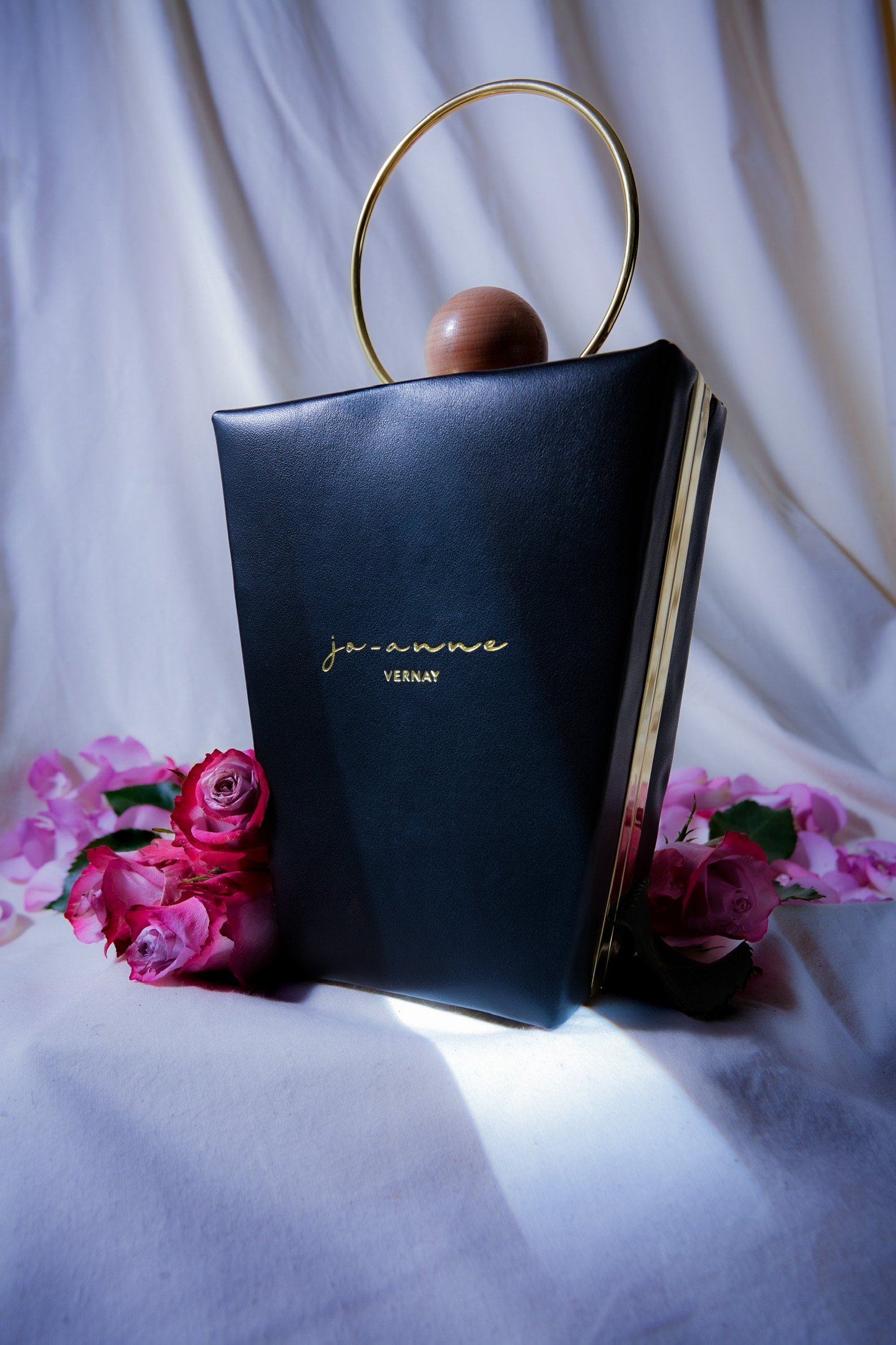 A black notebook with gold text that reads 'ja-anne VERNY' sits among pink roses and is topped with a round wooden ball encircled by a gold ring, all set against a draped white fabric background.