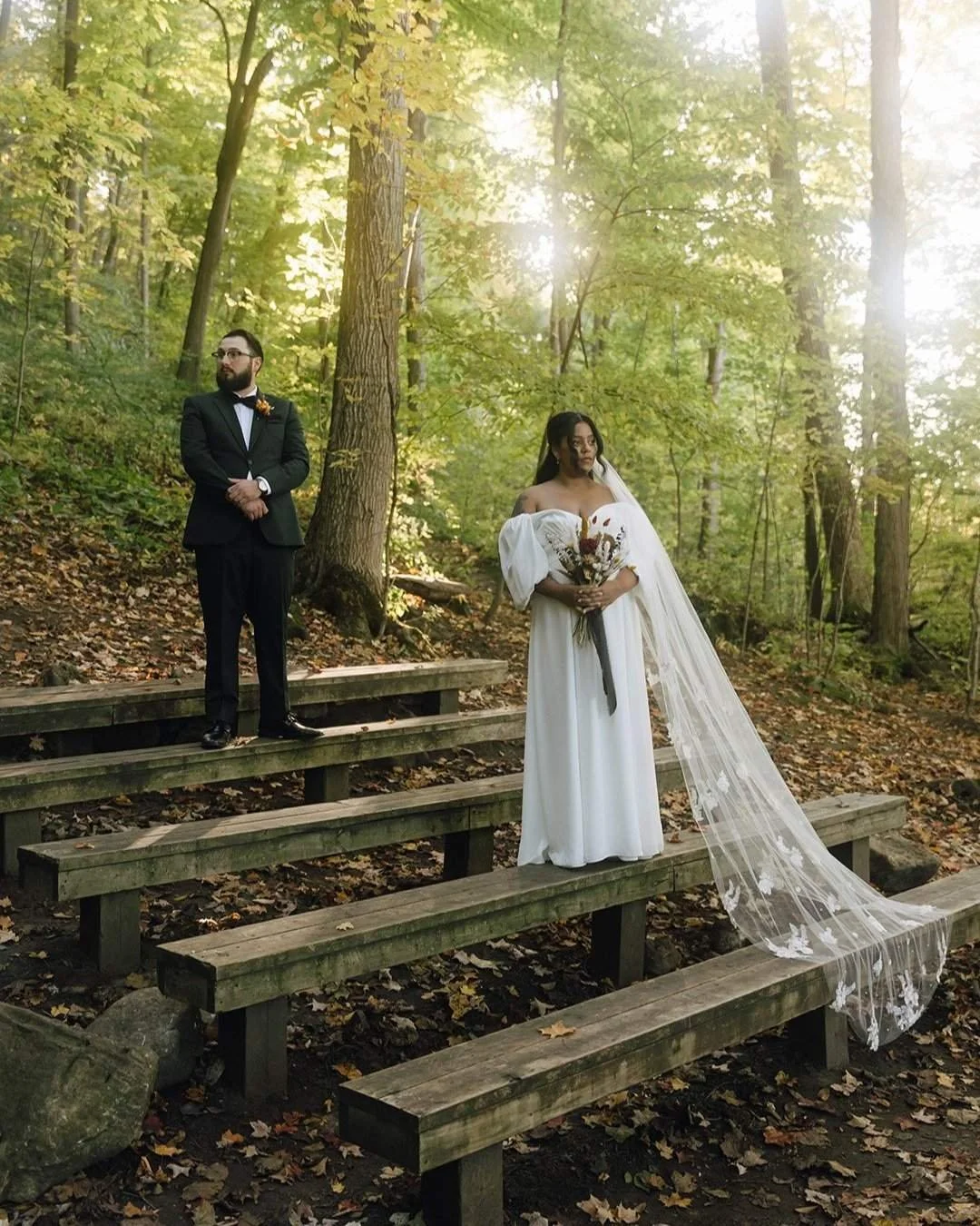 A bride and groom are posed, standing on bleachers in the forest looking opposite directions. Shot at Cave Springs Camp, Beamsville, Ontario