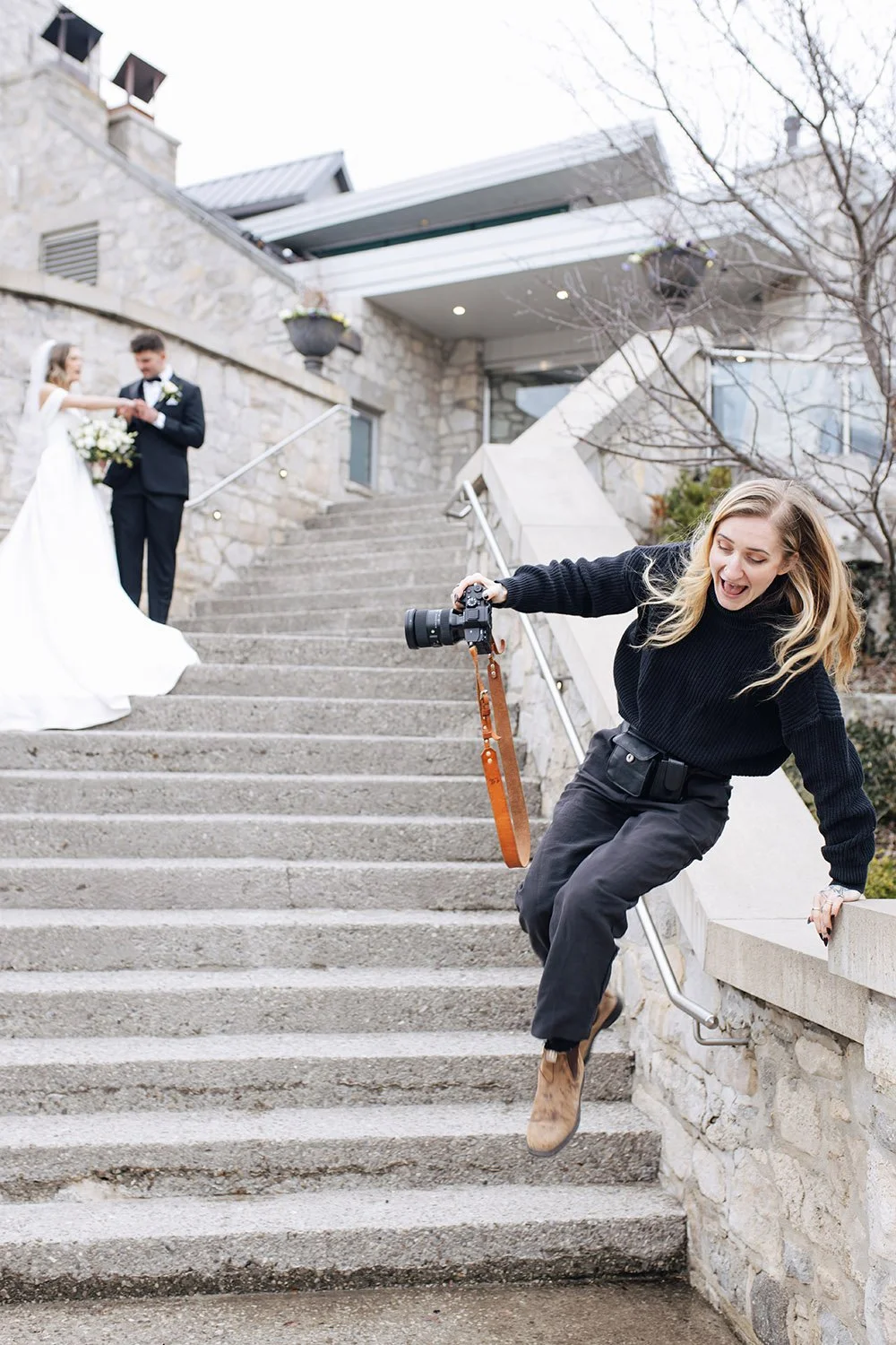 A wedding photographer captures a shot of a bride and groom standing on outdoor stairs. The photographer, a woman with blonde hair wearing a black sweater and dark pants, jumps off the railing with a camera in hand.