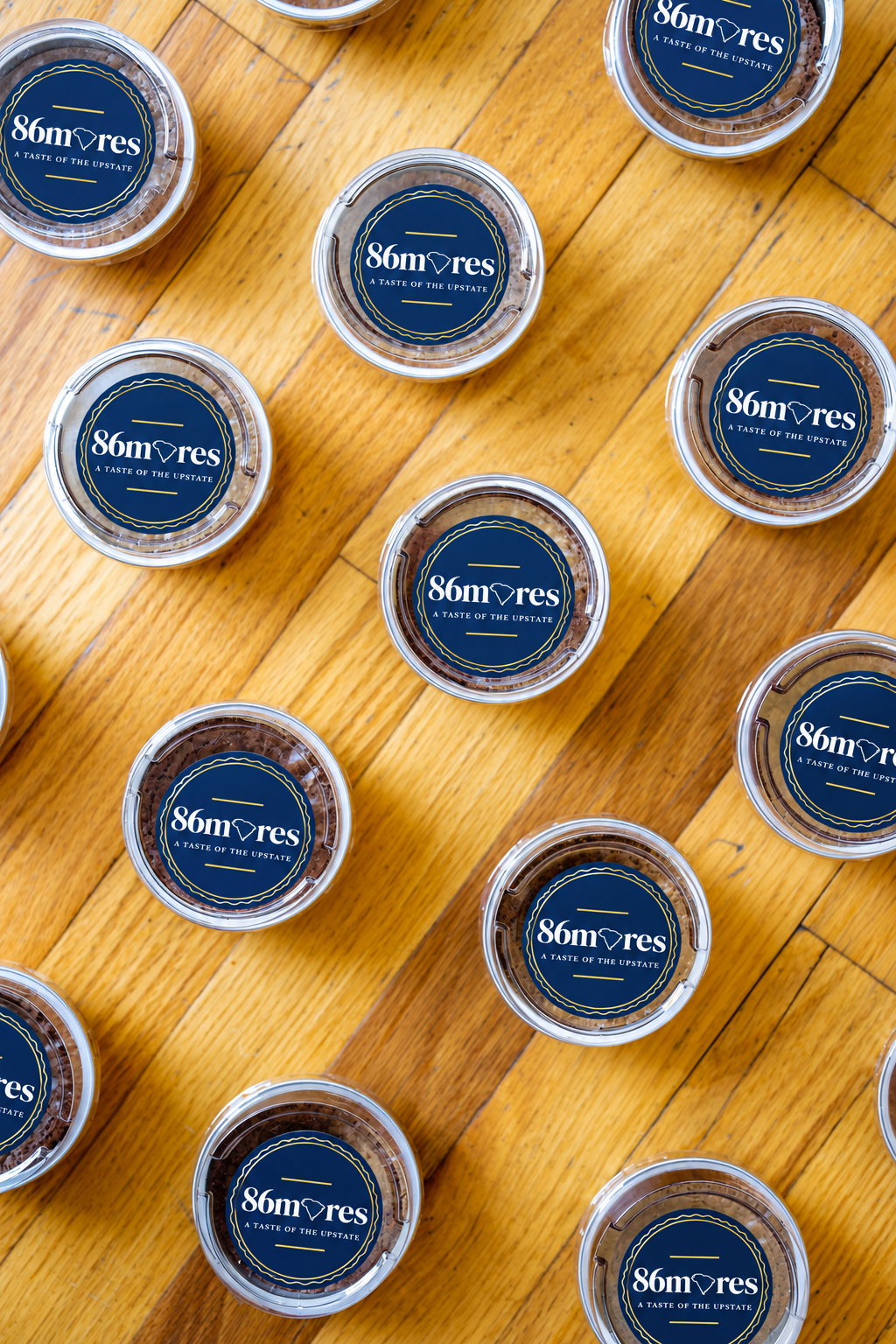 Top-down view of multiple small round containers with dark blue labels that read '86mores A Taste of the Upstate' on a wooden surface.