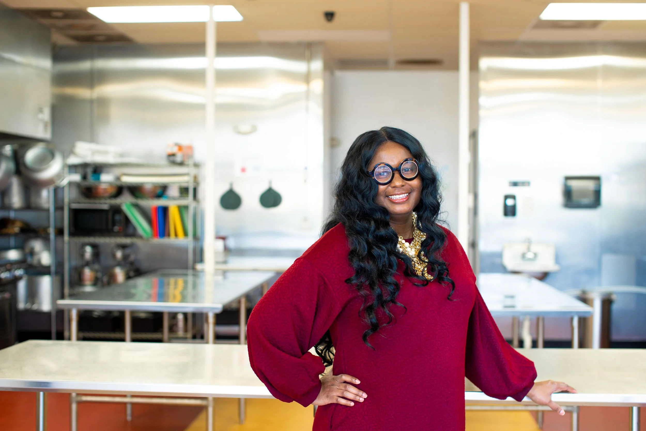 A woman with long, curly black hair, wearing large round glasses, a red sweater, and a statement necklace, standing in a professional kitchen.