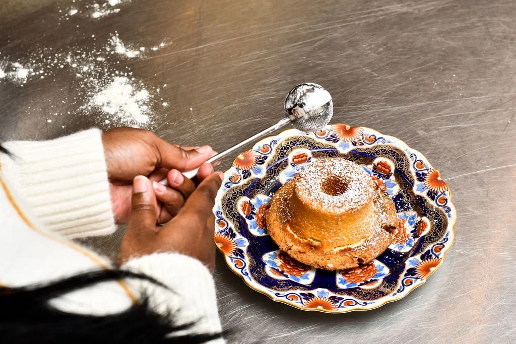 Person sprinkling powdered sugar on a bundt cake on a decorative plate.
