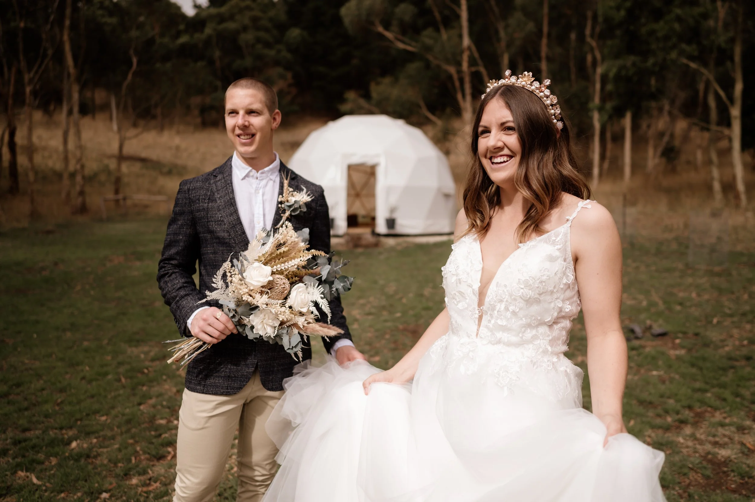 A bride and groom smiling outdoors during their wedding, with a white geodesic tent in the background.