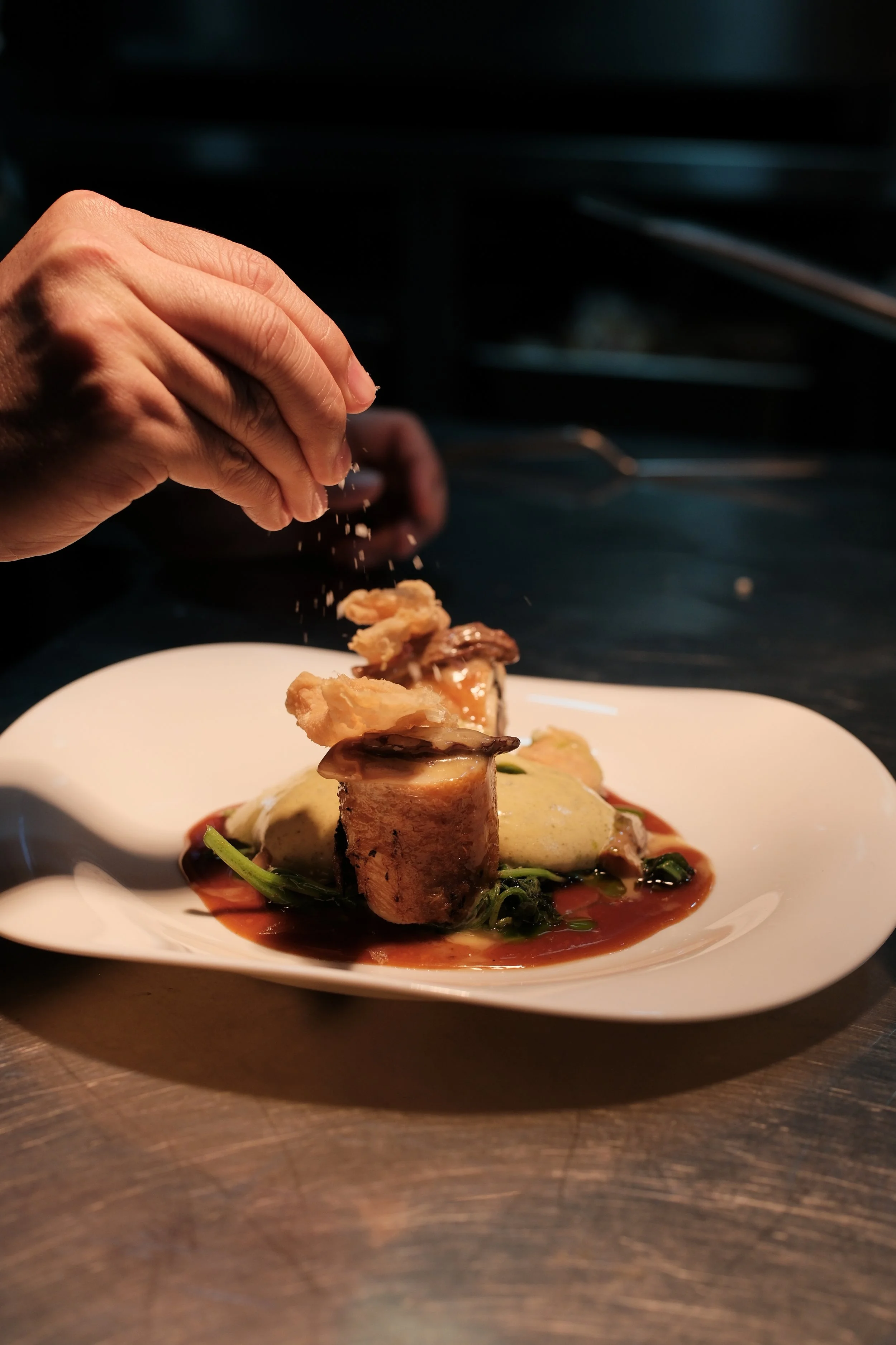 Chef garnishing a plated gourmet dish with seasoning, featuring steak, sauce, vegetables, and crispy toppings on a white plate in a professional kitchen.