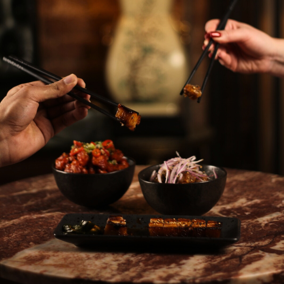 Two people using chopsticks to share a piece of grilled eel over a table with Asian dishes, including bowls of spicy tuna poke and shredded cabbage salad.