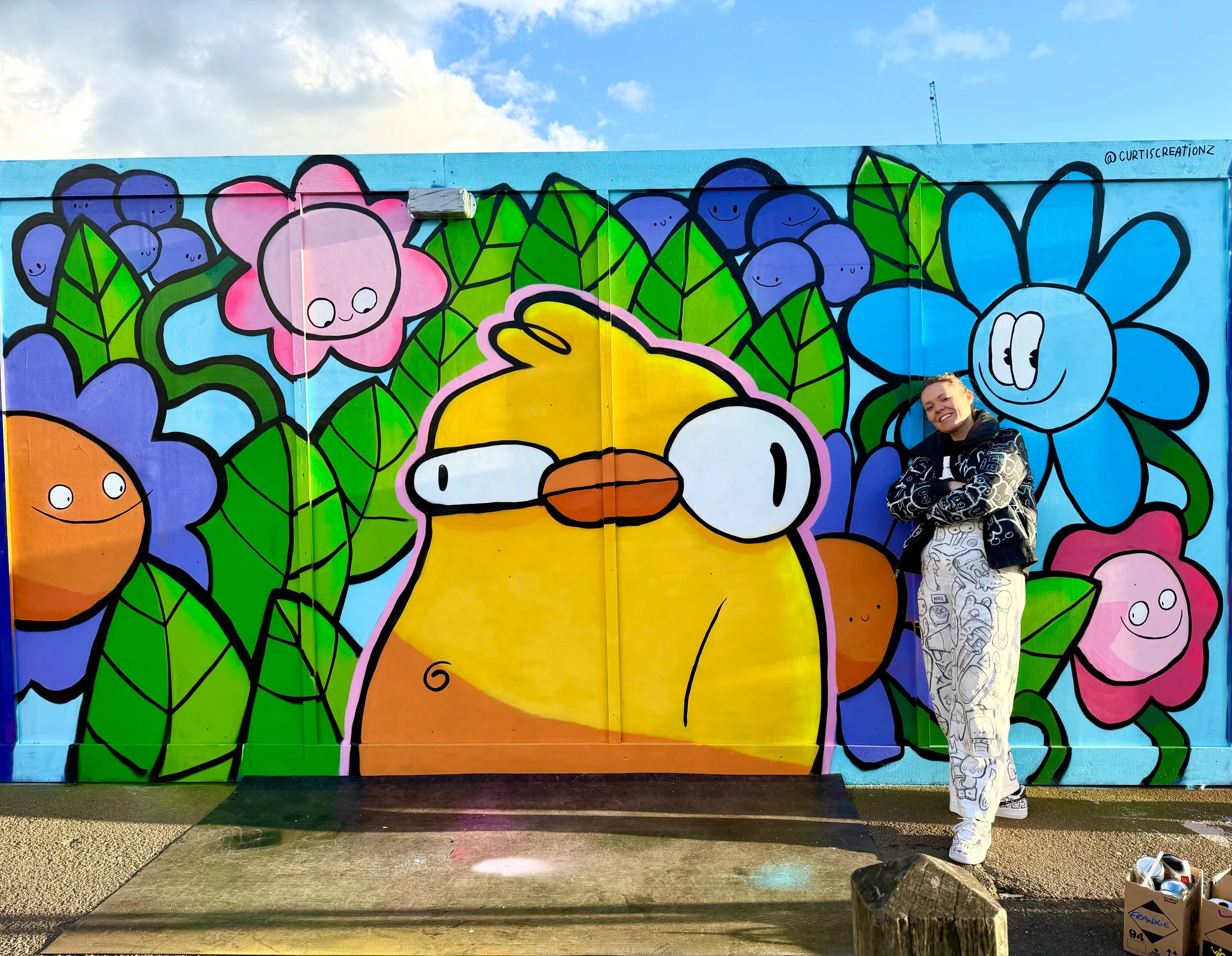 Colorful mural with cartoon-style flowers and leaves, and a smiling person standing in front of it. The mural is painted on a wooden fence with a blue sky and clouds above.