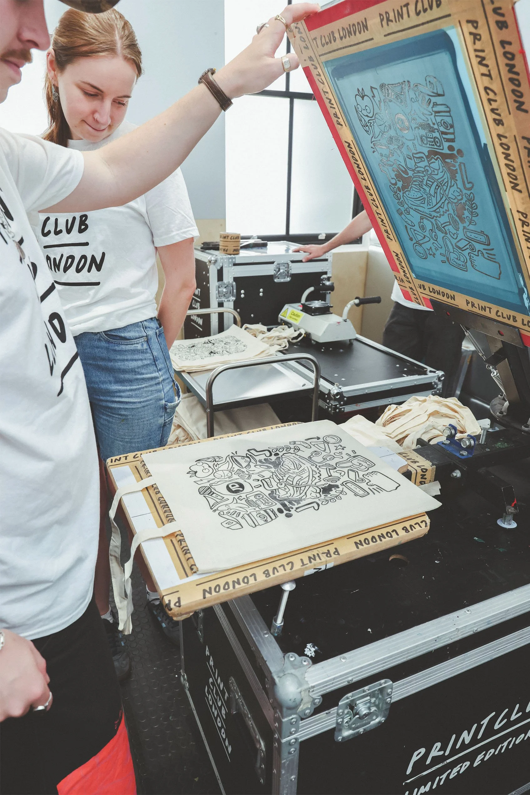 A group of people observing a large screen or printer printing a detailed, black-and-white artistic design on fabric bags at a printmaking event or workshop.