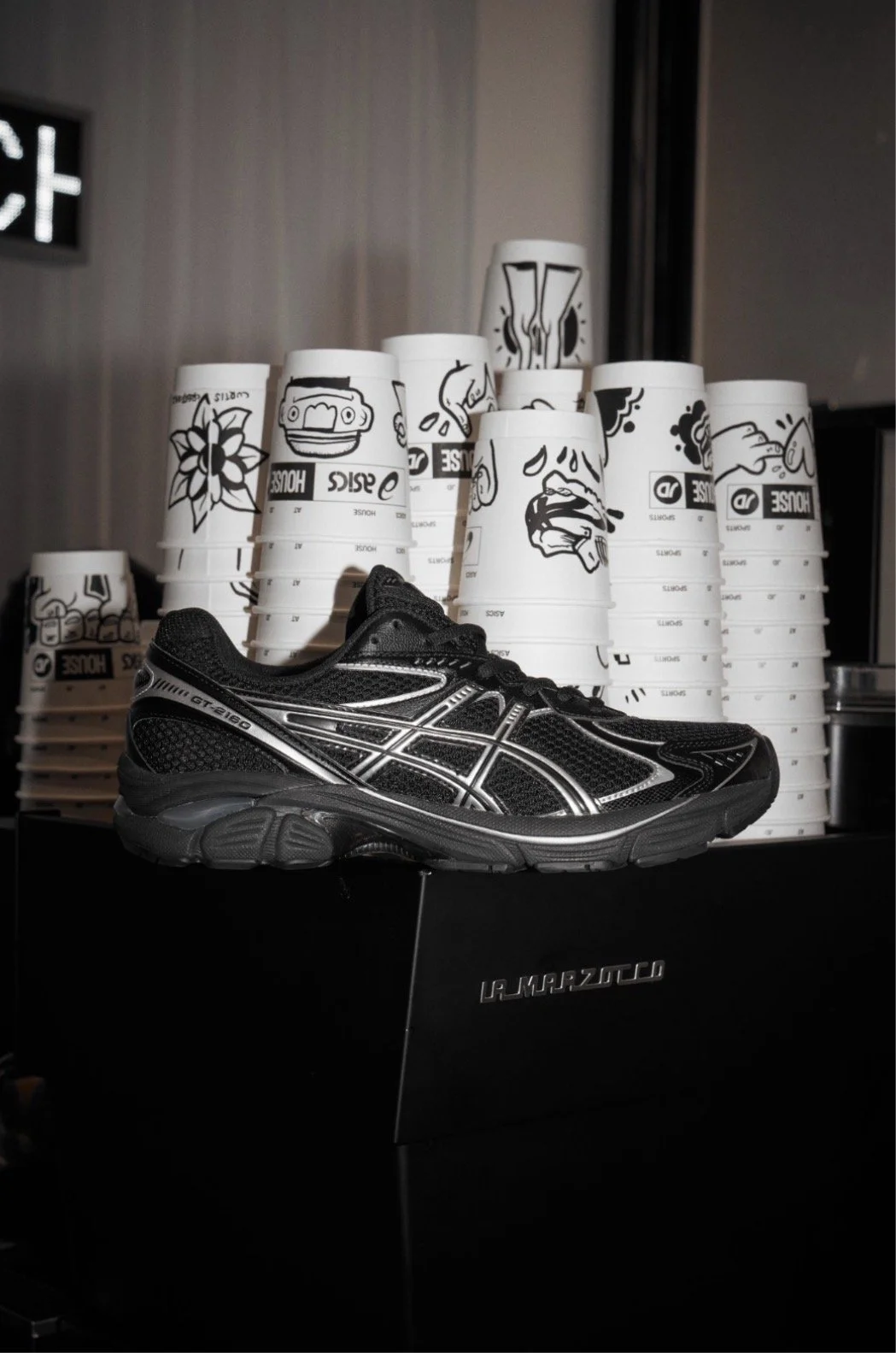 A black athletic shoe displayed on a black box, with stacks of white paper cups featuring black cartoon-style drawings and text in the background.