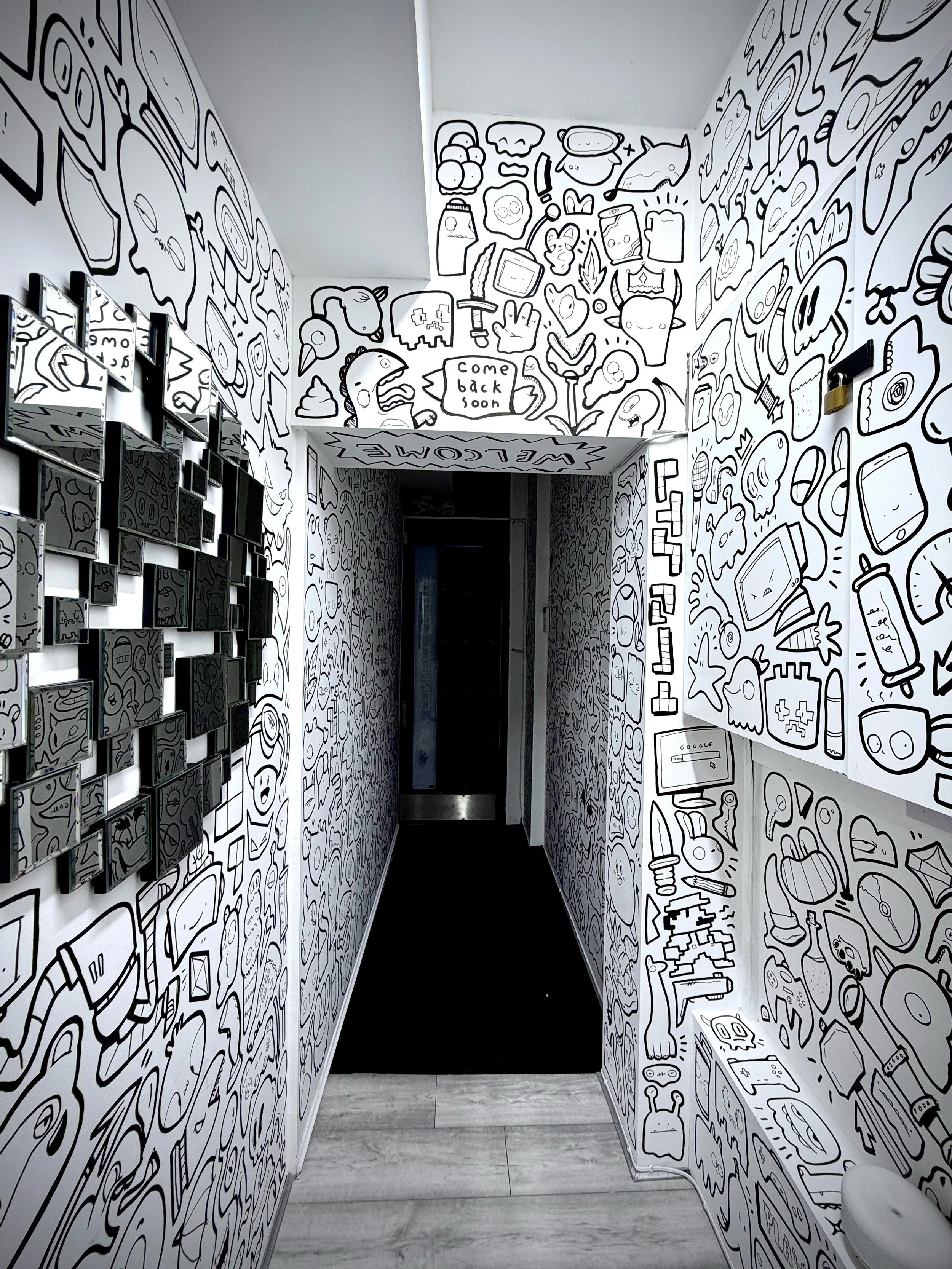 Black and white doodle art on the walls of a narrow hallway, featuring various cartoonish characters, objects, and symbols, including a fish, a sword, a hand making a peace sign, and a speech bubble saying 'Come back soon.'