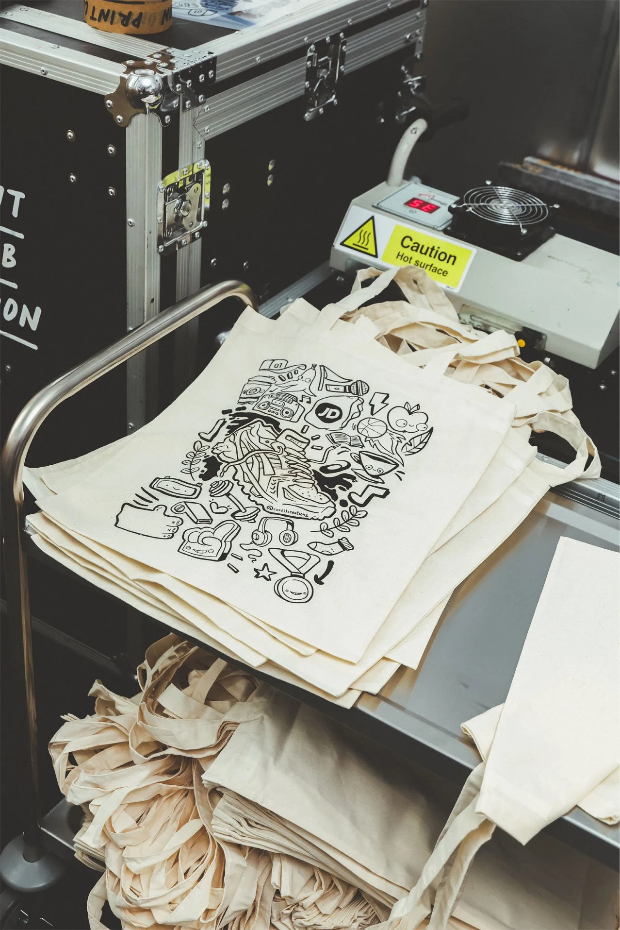 A cart holding multiple stacks of beige tote bags with black graphic designs, and some bags are scattered underneath the cart. The cart is near a machine with a yellow caution sign indicating a hot surface, and a digital display showing the number 36